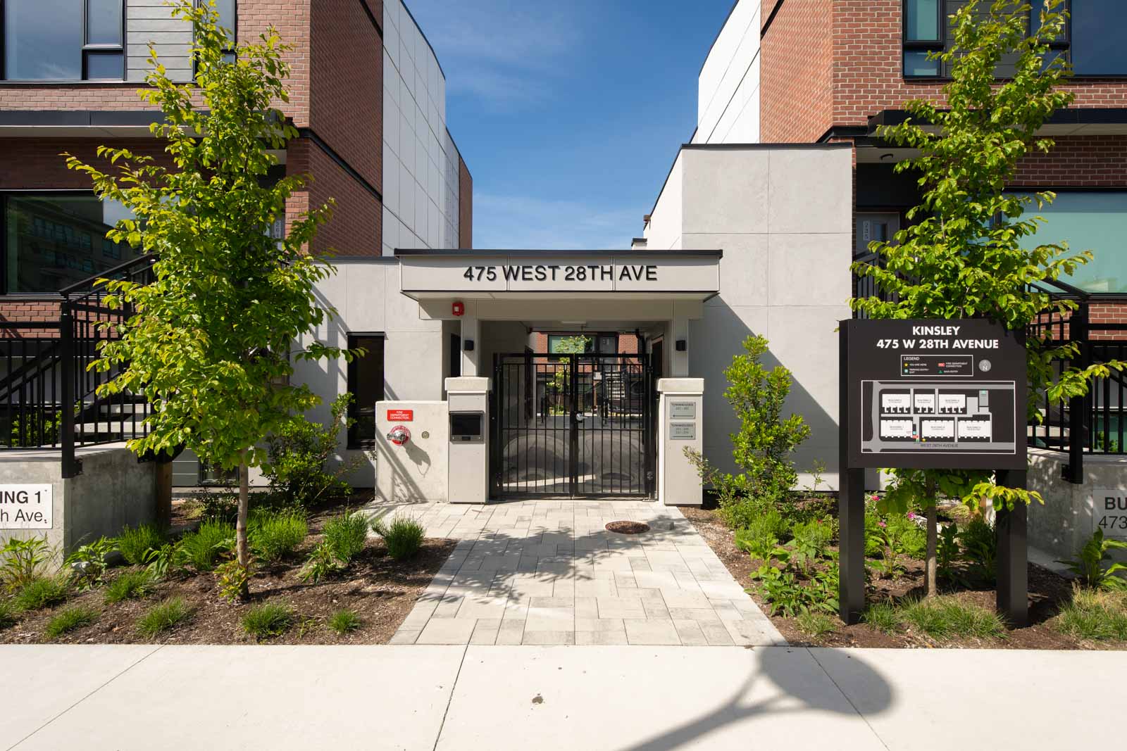 Entrance to a modern apartment building at 475 West 28th Ave with a secure gate, sign displaying address, and landscaped walkway with young trees.