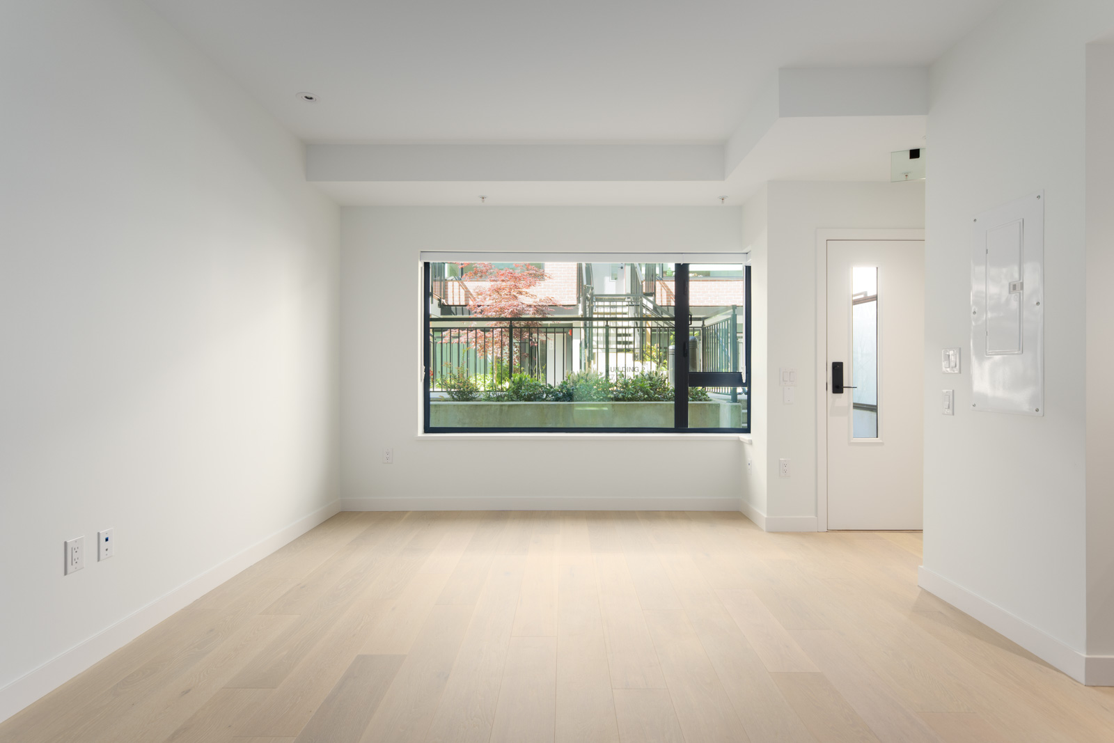 Empty, unfurnished room with light wood flooring, white walls, a large window, and a door. Natural light comes in through the window, showing an outdoor area with plants.