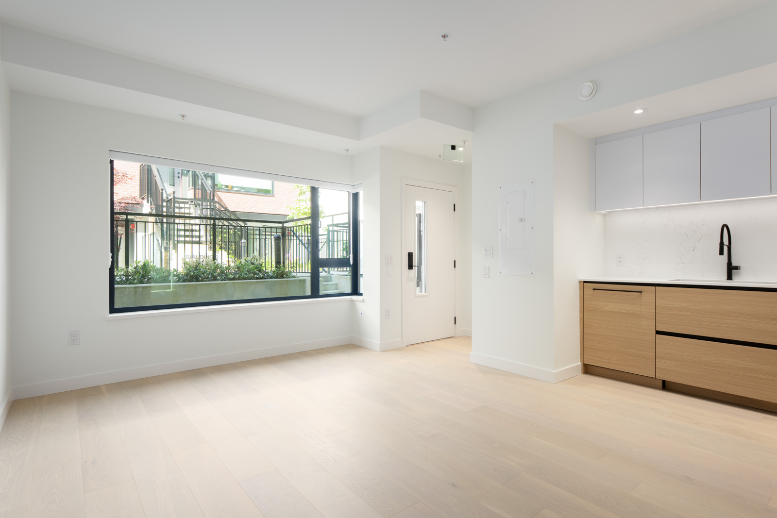Bright, unfurnished modern apartment with large window, light wood floors, white walls, and a minimalist kitchen featuring wood lower cabinets and white upper cabinets.