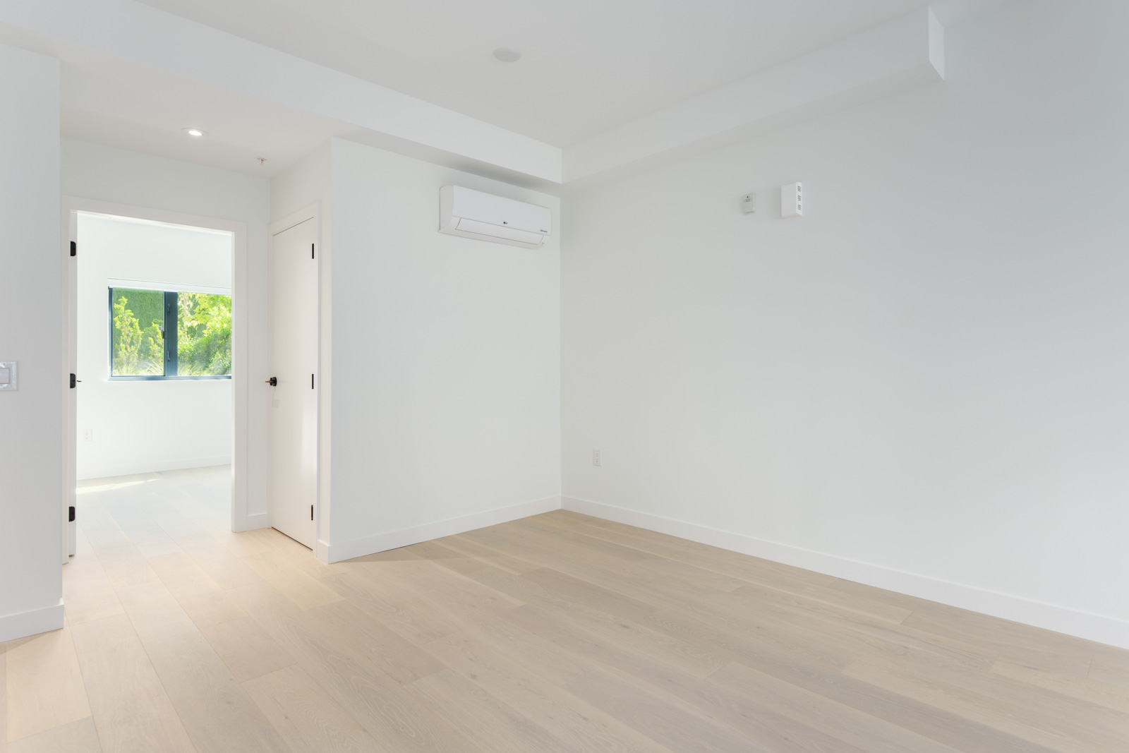Empty, unfurnished room with light wood floors, white walls, and an air conditioning unit; a doorway leads to another room with a window showing greenery outside.