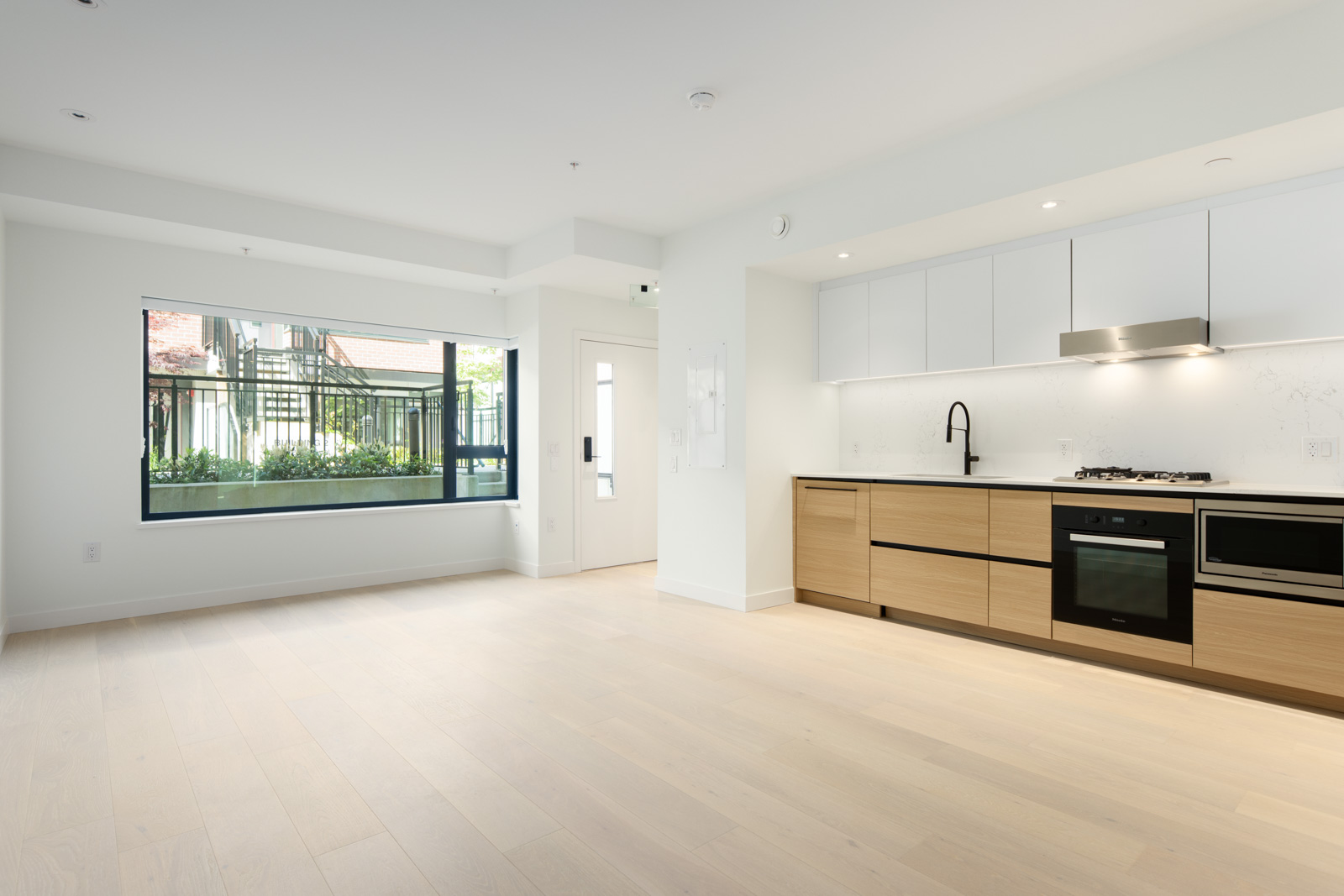 Modern, unfurnished apartment kitchen with light wood floors, white cabinets, built-in oven, stove, and a large window providing natural light.