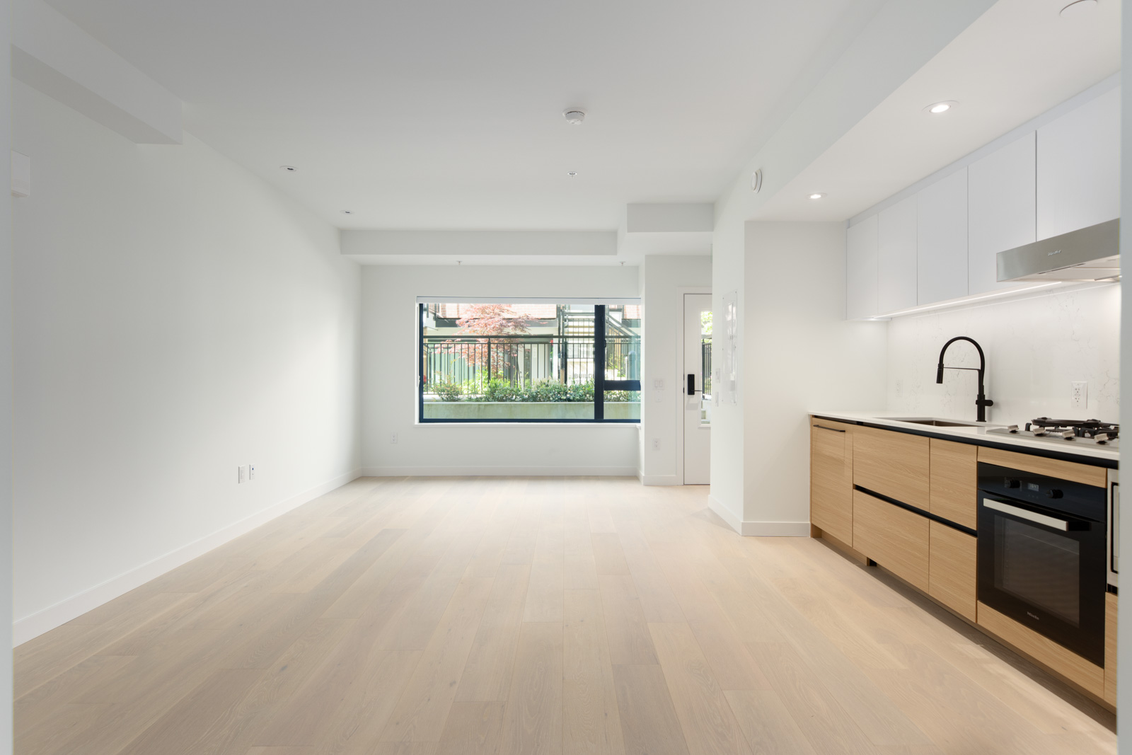Empty modern apartment with light wood floors, white walls, large window, and an open kitchen featuring light wood and white cabinetry with a black faucet and built-in appliances.
