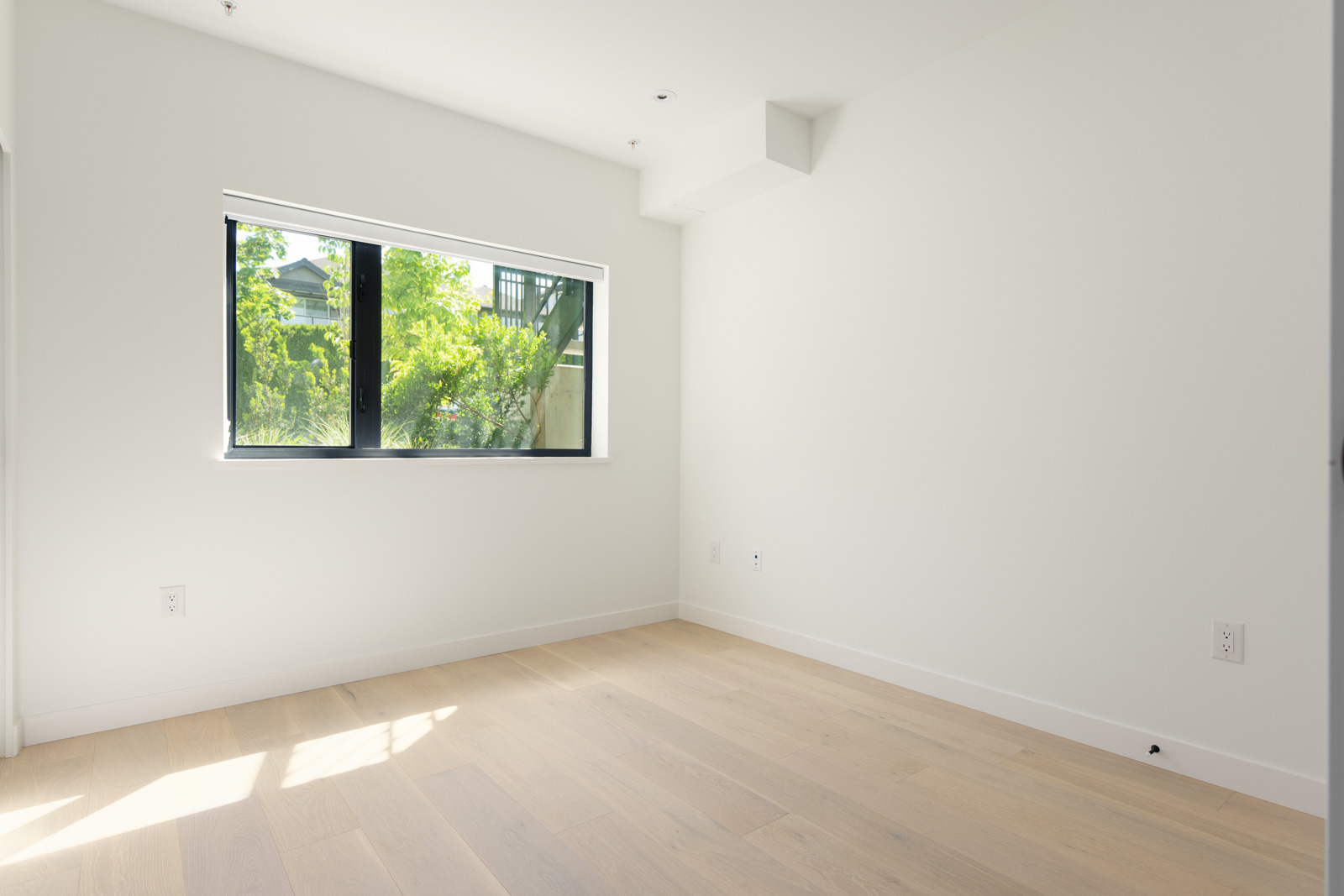 Minimalist, empty room with light wood flooring, white walls, and a large window letting in natural light and showing greenery outside.