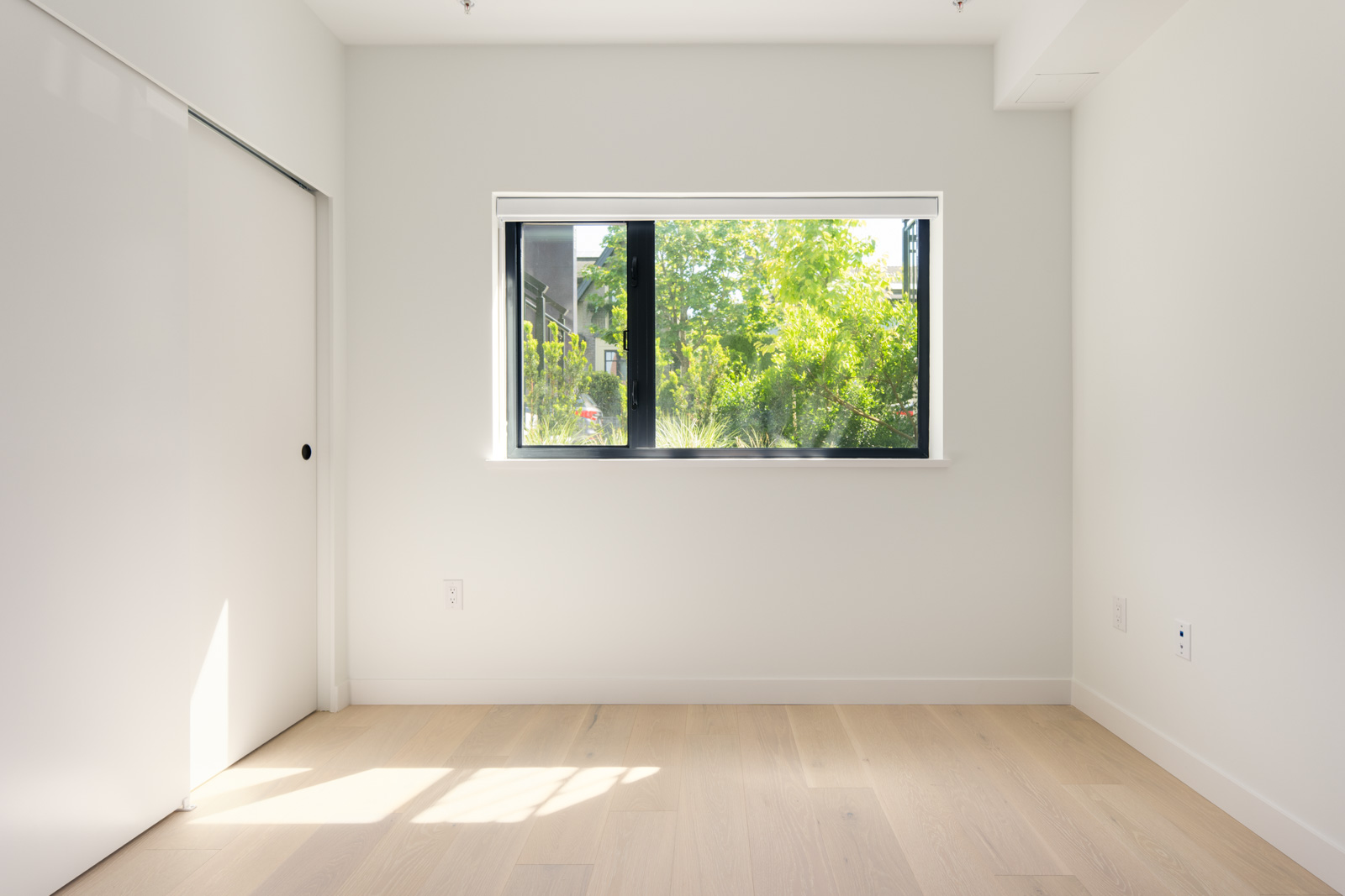 Empty room with light wood flooring, white walls, a large window showing green trees outside, and a closed white sliding door on the left.