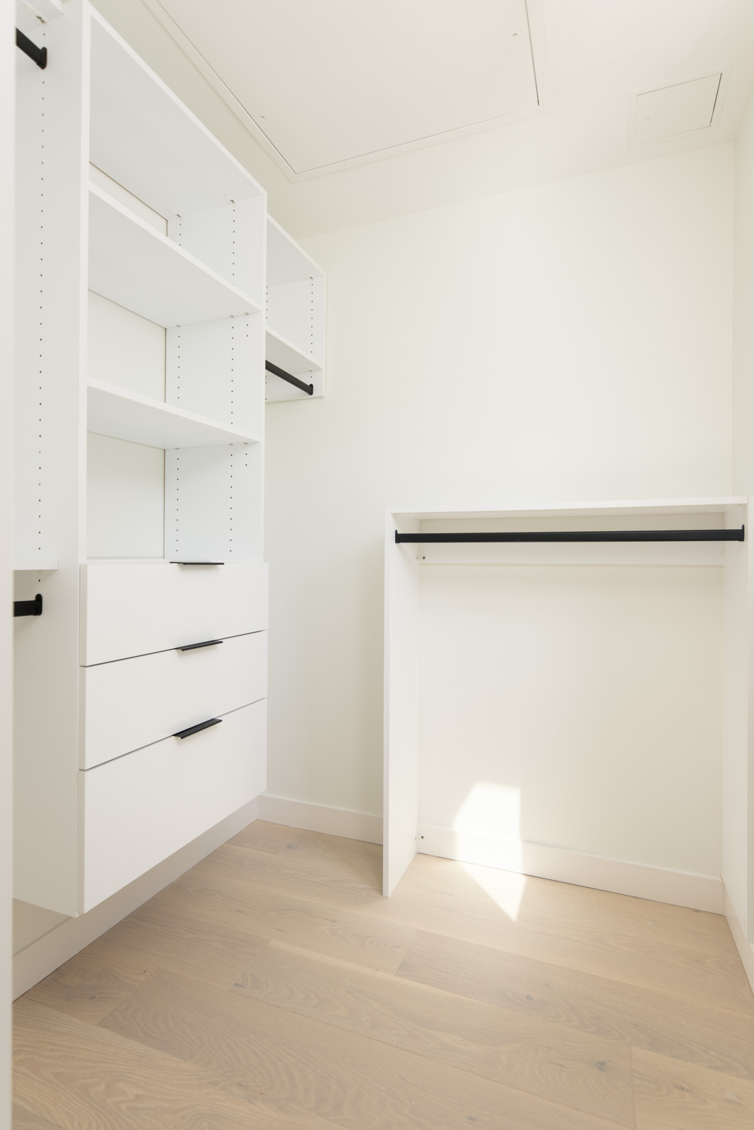 Minimalist walk-in closet with white shelving, drawers, and an empty hanging rod, featuring light wood flooring and plain white walls. Sunlight is entering from the right side.