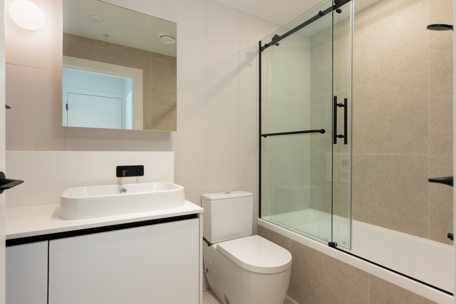 Modern bathroom with a sink, cabinet, wall mirror, toilet, and a bathtub with a glass shower screen and black fixtures. Neutral color scheme with beige and white tiles.