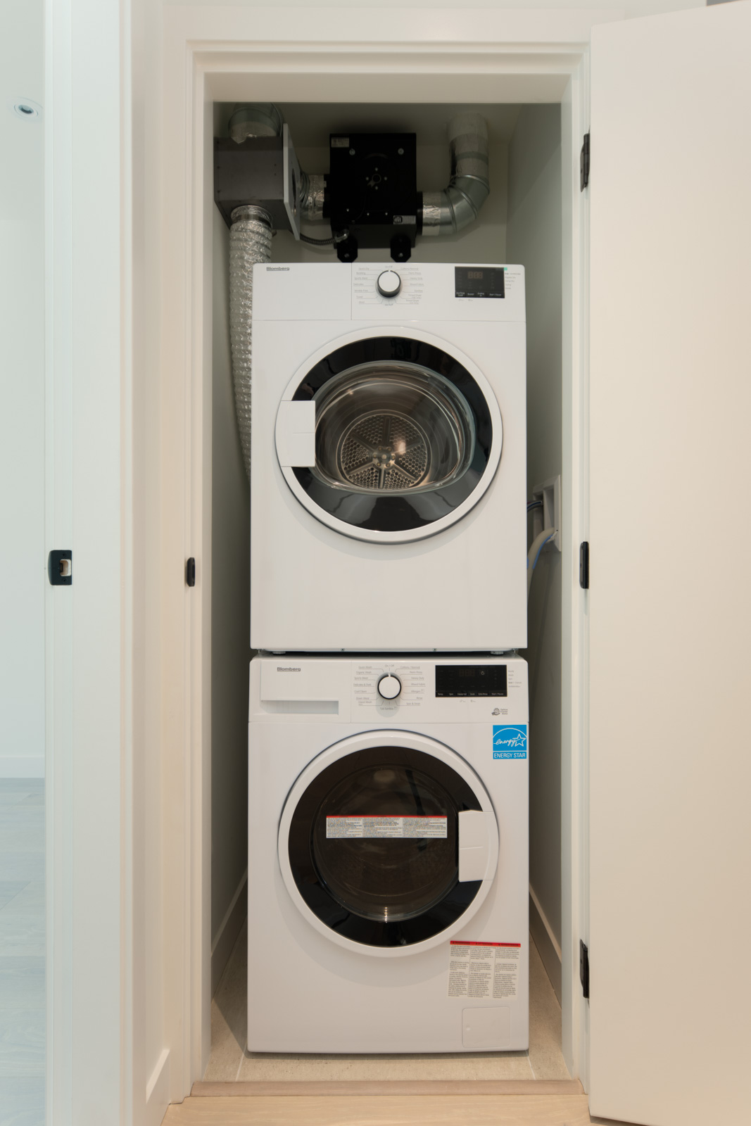 Stacked washer and dryer units are installed in a small laundry closet with white doors and venting system above.