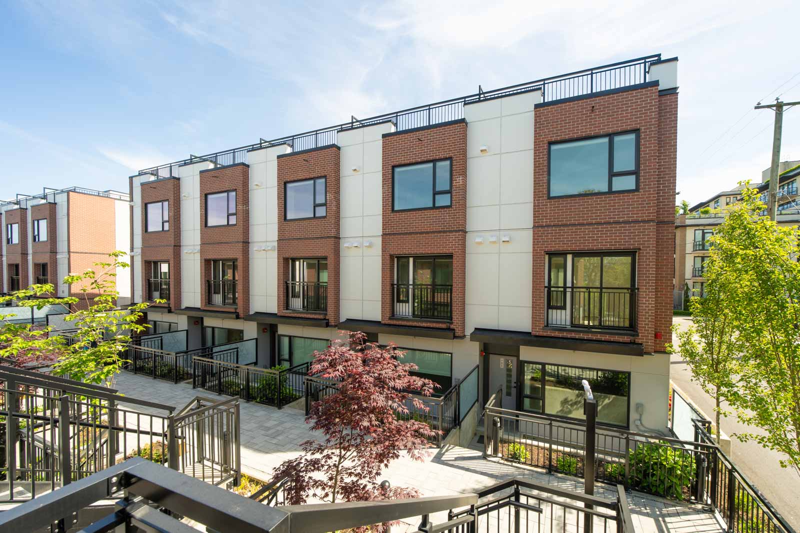A row of modern three-story townhouses with brick and white exteriors, large windows, and small balconies, surrounded by trees and metal railings.