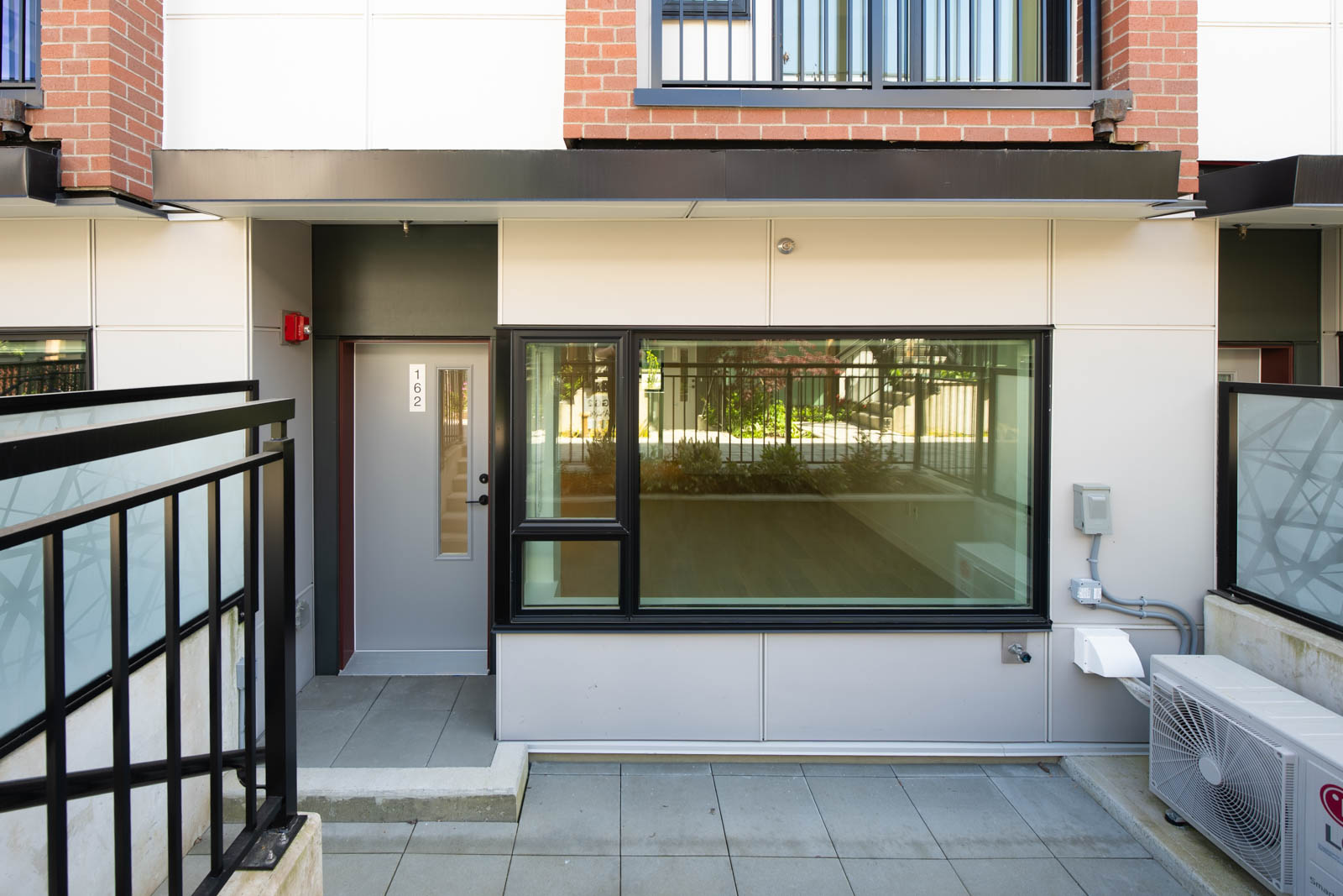 Ground-level view of a modern townhouse entrance with a large window, a black metal railing, and an outdoor air conditioning unit.