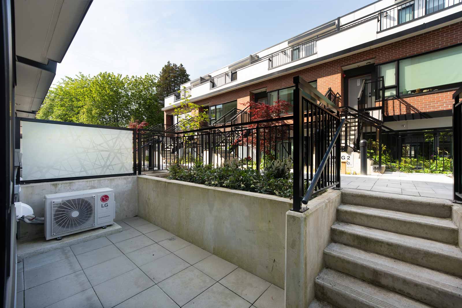 Ground-level patio with tiled flooring, a railing, stairs leading up, an air conditioning unit, and a view of modern townhouse buildings with greenery.
