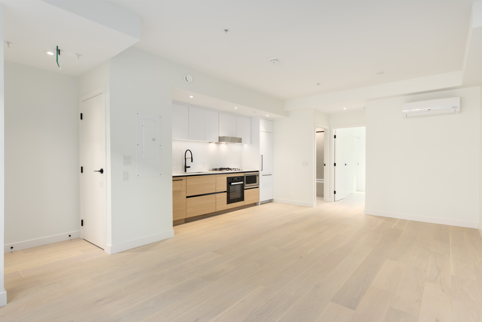 Modern, unfurnished apartment with light wood flooring, white walls, an open kitchen area with built-in appliances, and multiple doors leading to other rooms.