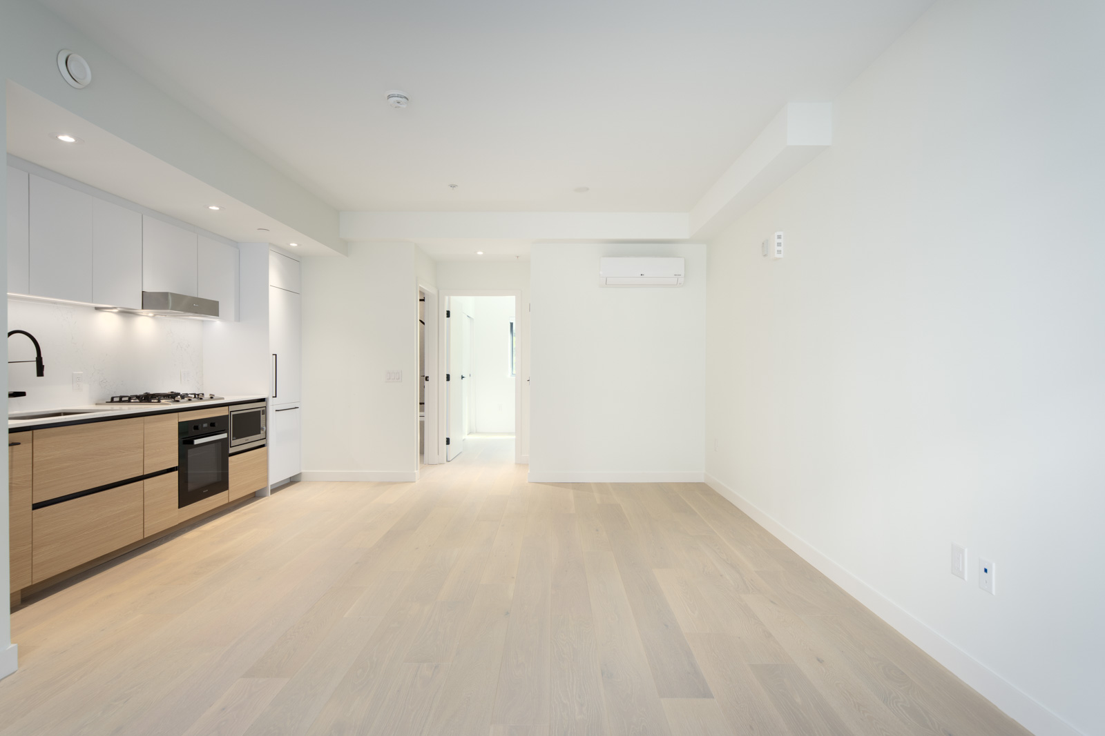 Modern, unfurnished apartment with light wood floors, white walls, built-in kitchen appliances, and recessed lighting, featuring an open layout and natural light from a distant window.