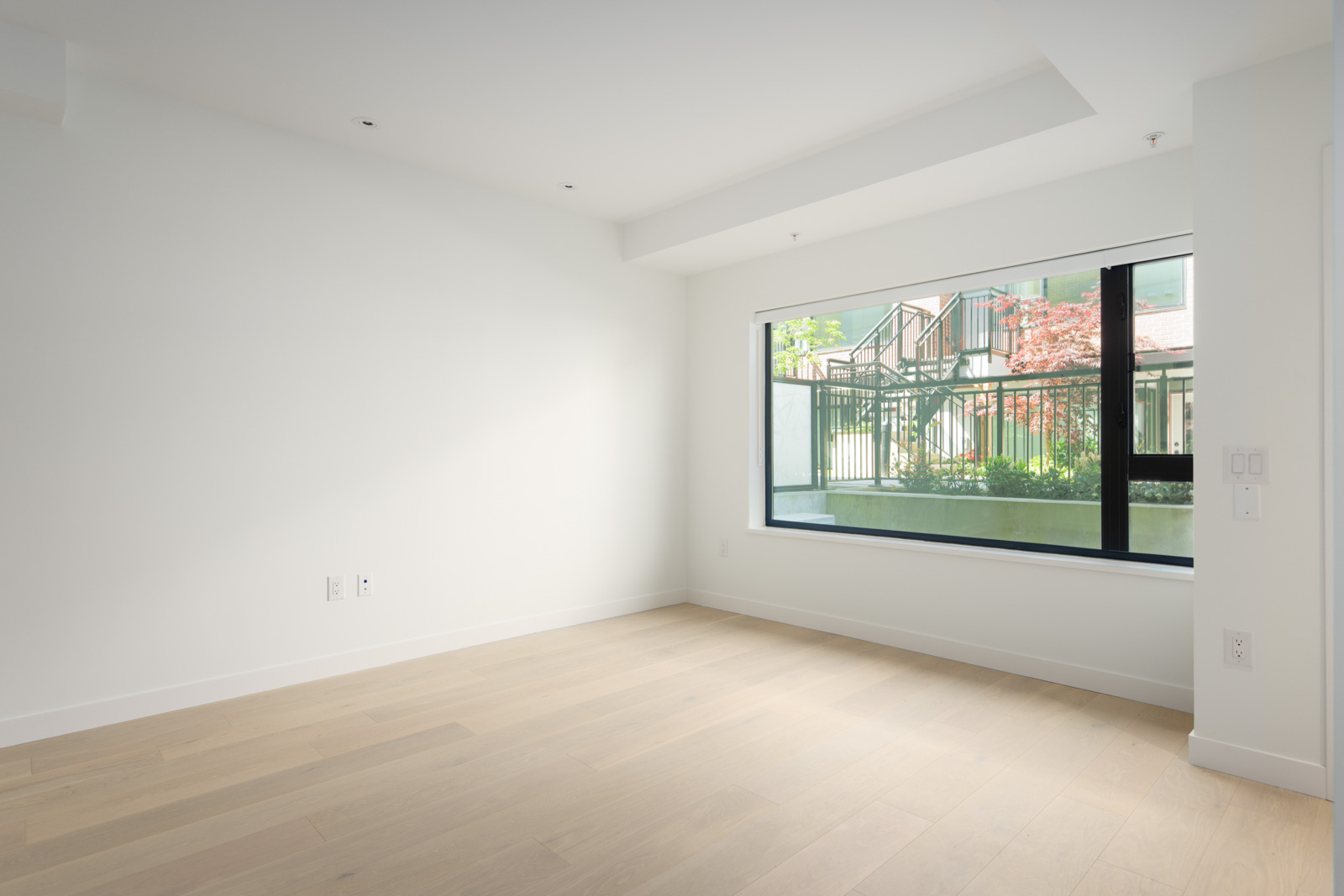 Empty room with white walls, light wood flooring, and a large window letting in natural light and showing an outdoor view of greenery and a building.