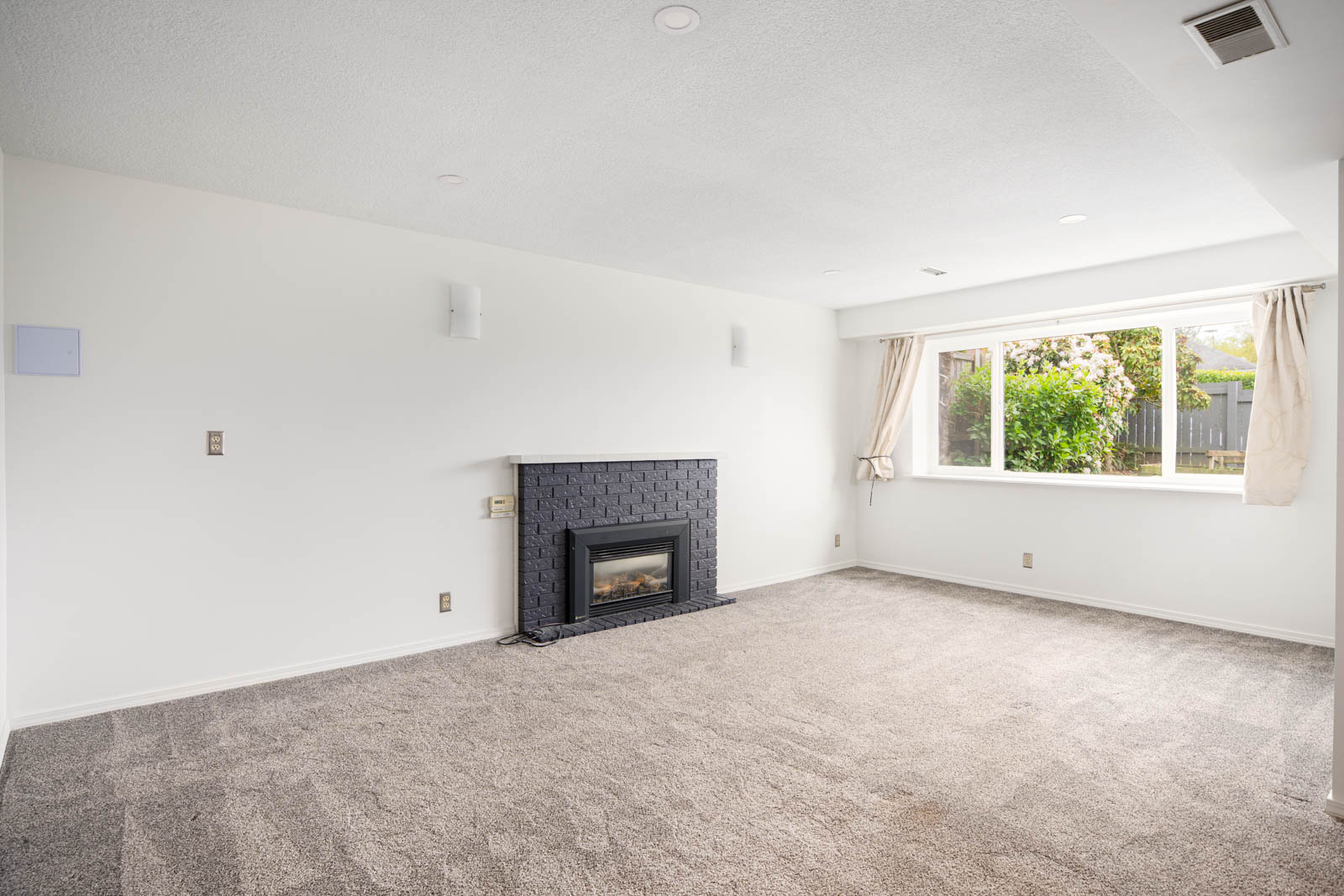 Unfurnished living room with gray carpet, white walls, a black tile fireplace, and a large window with open beige curtains facing a garden.