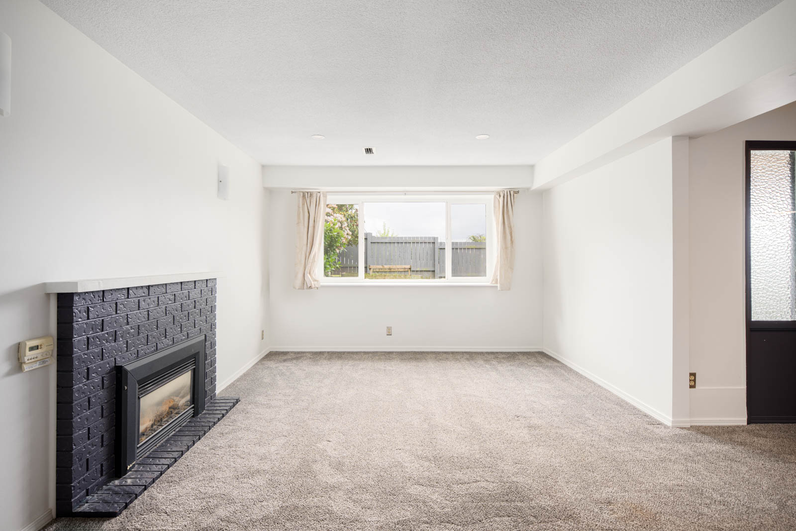 Unfurnished living room with a gray carpet, black brick fireplace, white walls, large window with curtains, and view of a fenced yard.