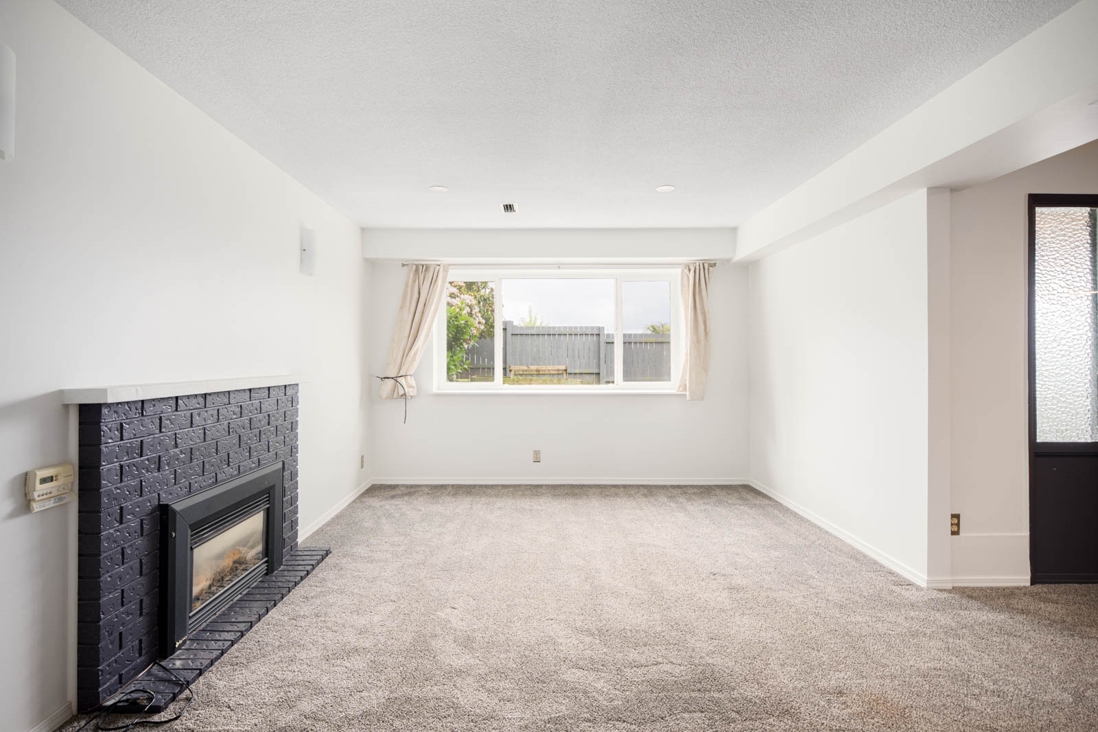 Unfurnished living room with grey carpet, white walls, a black brick fireplace, large window with curtains, and a frosted glass door.