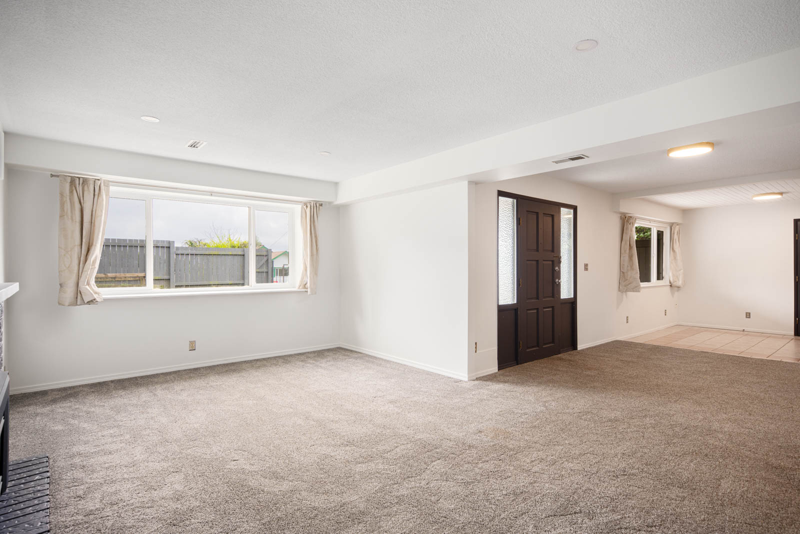 Unfurnished living room with beige carpet, white walls, large window with curtains, and a dark front door leading to a tiled entry area.