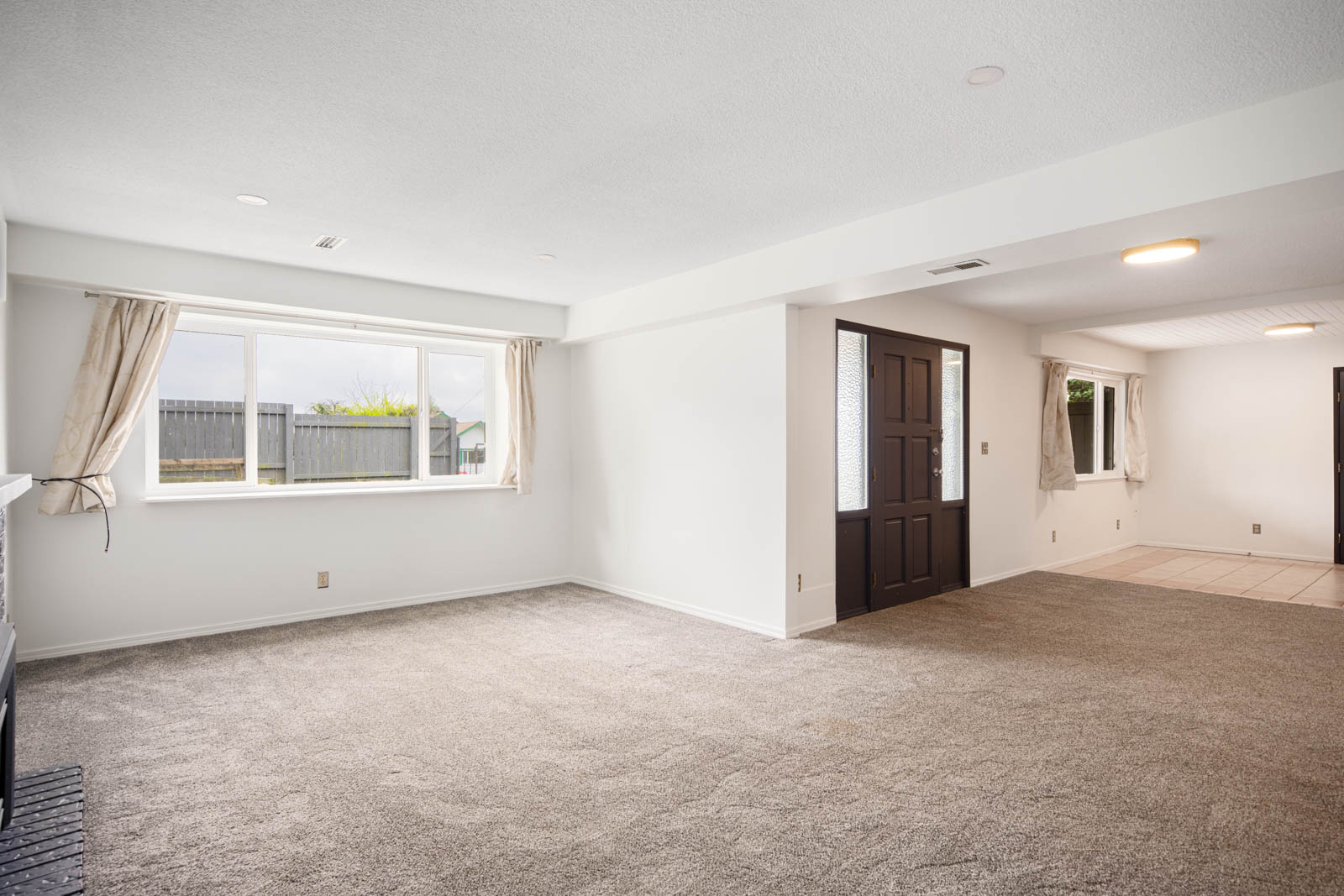 Spacious empty room with beige carpet, white walls, large window with curtains, and a dark front door leading to a tiled entryway.