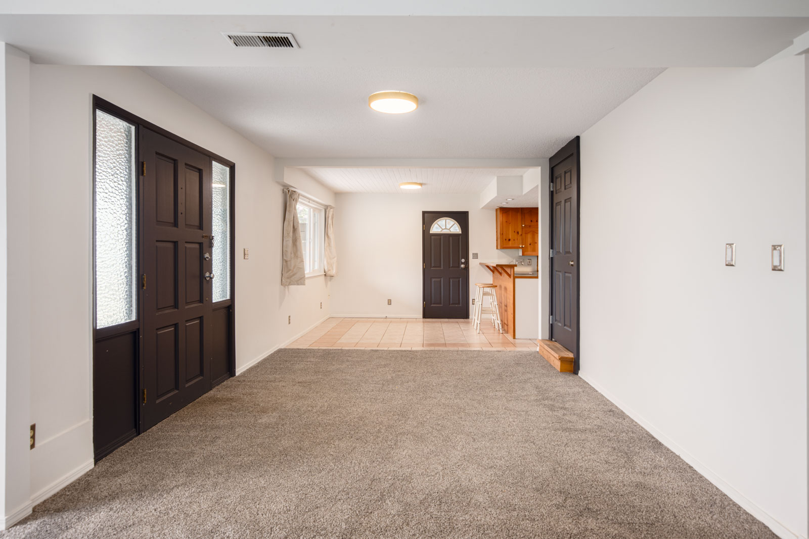 A room with gray carpet, white walls, black entry doors, and a tiled area leading to a small kitchen with wood cabinets.