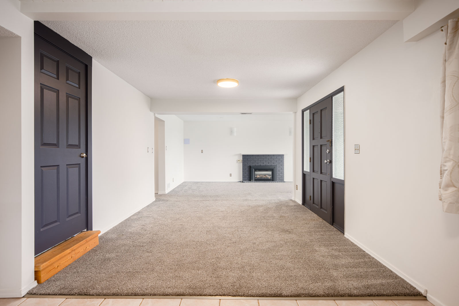 Spacious, carpeted living room with a fireplace, white walls, dark doors, and a ceiling light fixture.