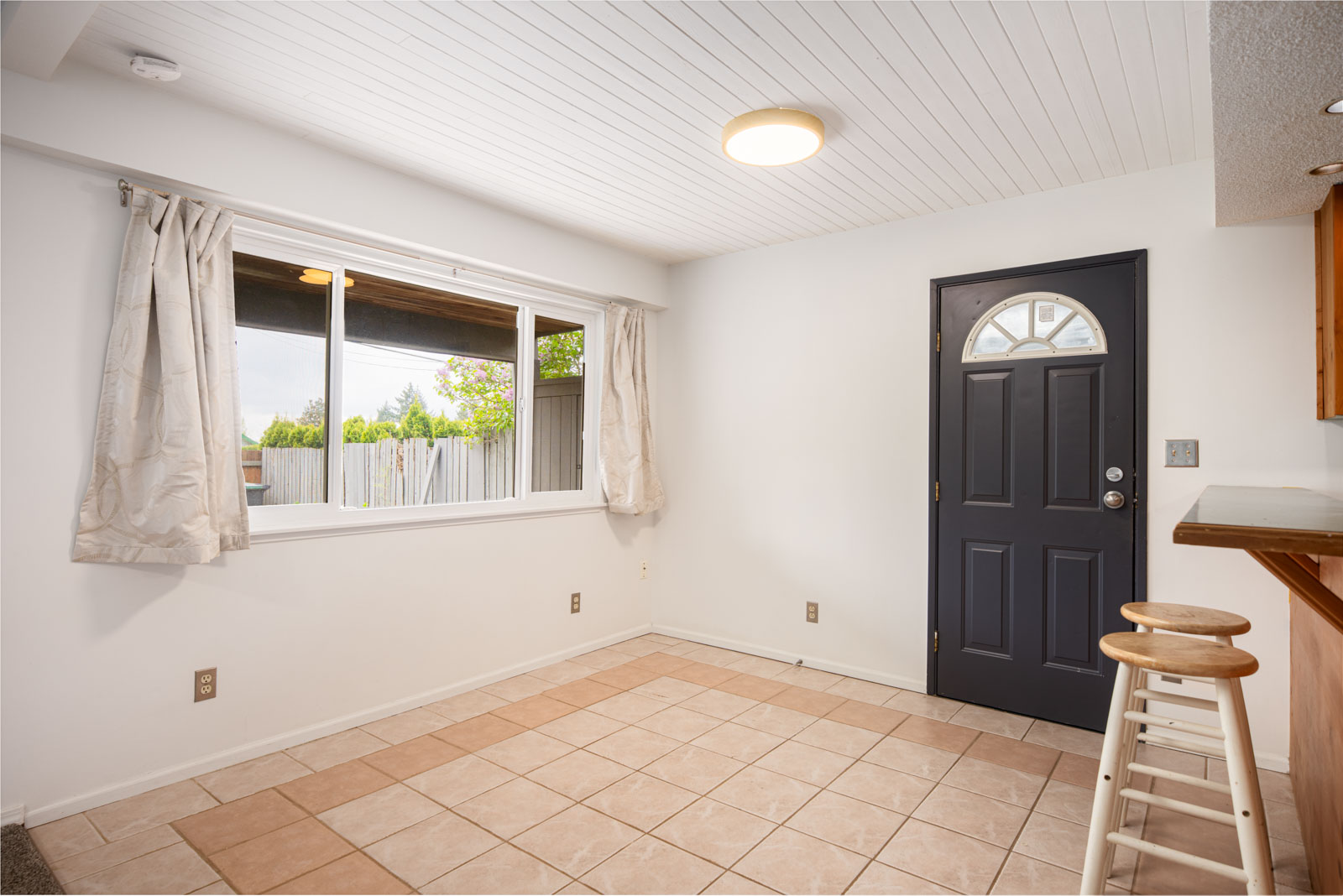 A small empty room with tiled floor, white walls, a window with curtains, a black front door, and two wooden stools by a counter.