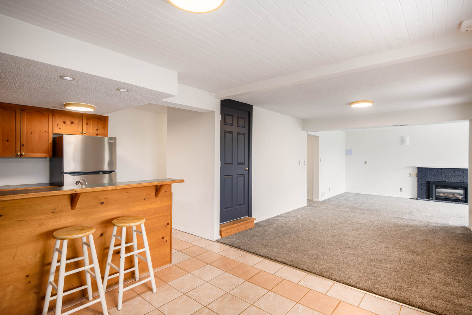 Open basement area with tiled flooring near a wood-paneled kitchen, carpeting in the living space, a dark door, and a fireplace in the corner. Two stools are at the kitchen counter.