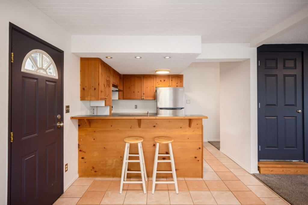 Small kitchen with wooden cabinets and countertop, two stools at the bar, stainless steel fridge, tile floor, and two dark-colored doors.