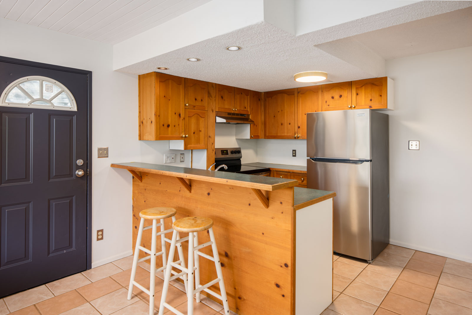 Compact kitchen with wood cabinets and breakfast bar, two wooden stools, stainless steel appliances, and a dark front door; tile flooring throughout.