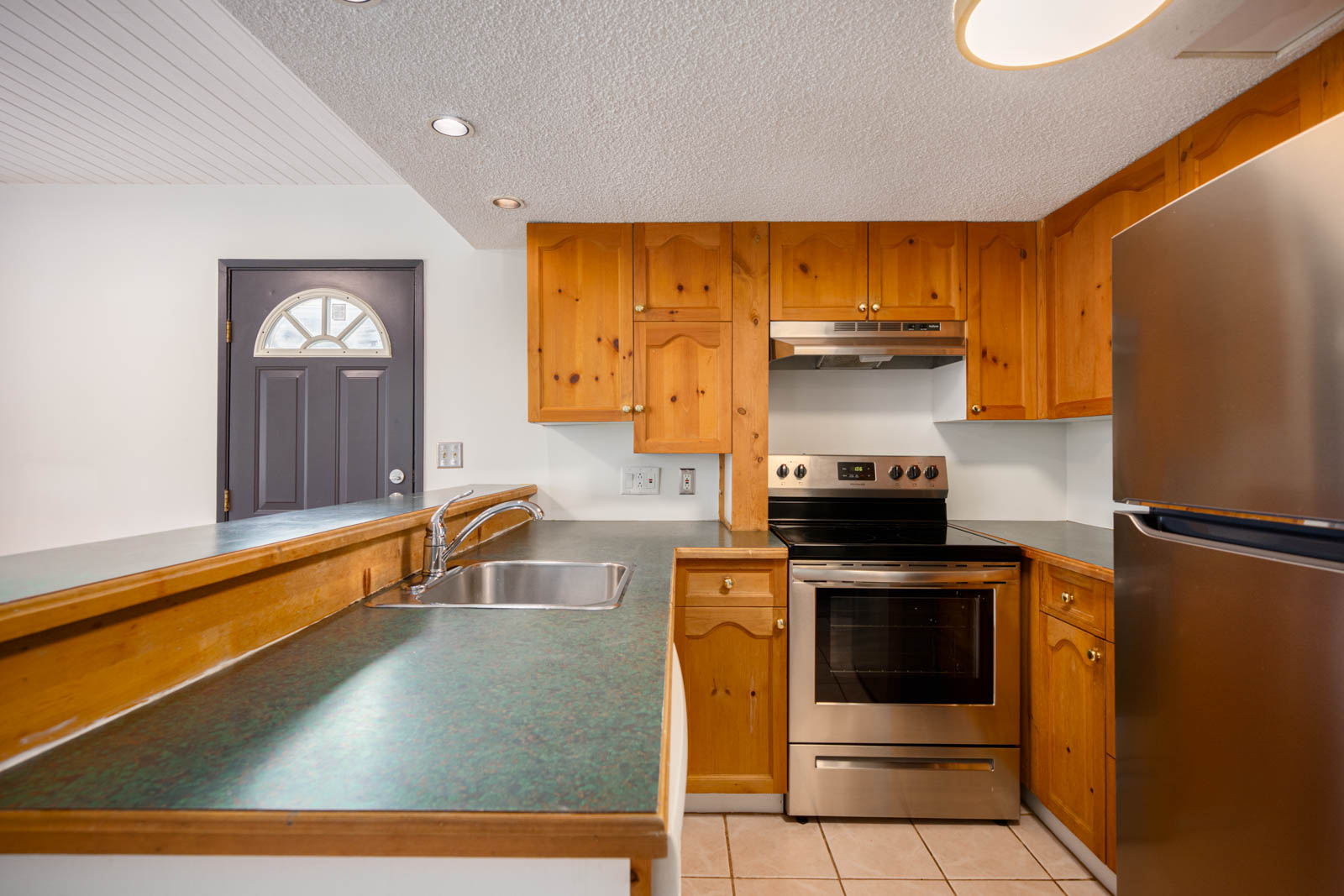 Kitchen with wooden cabinets, stainless steel appliances, a double sink, green countertops, and a gray door in the background.