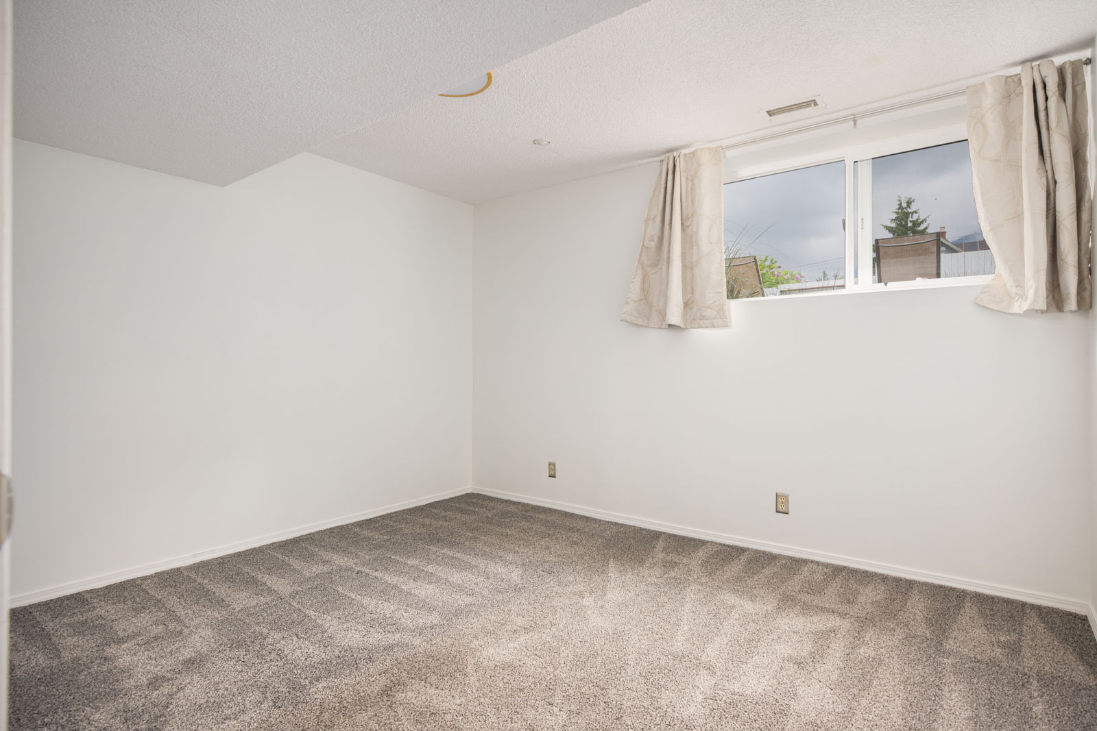 Empty room with white walls, a small window with beige curtains, and carpeted flooring; no furniture or decorations are present.