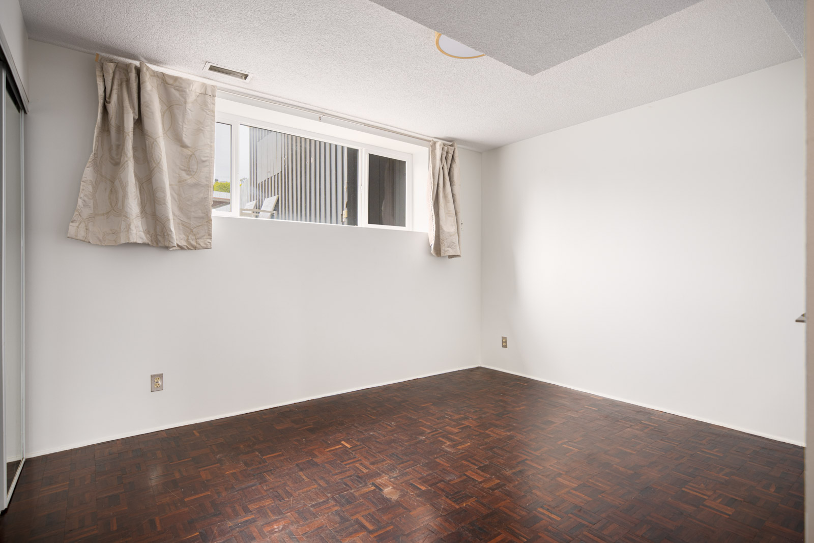 Empty room with white walls, a parquet wood floor, a large rectangular window with curtains, and a mirrored closet door on the left.