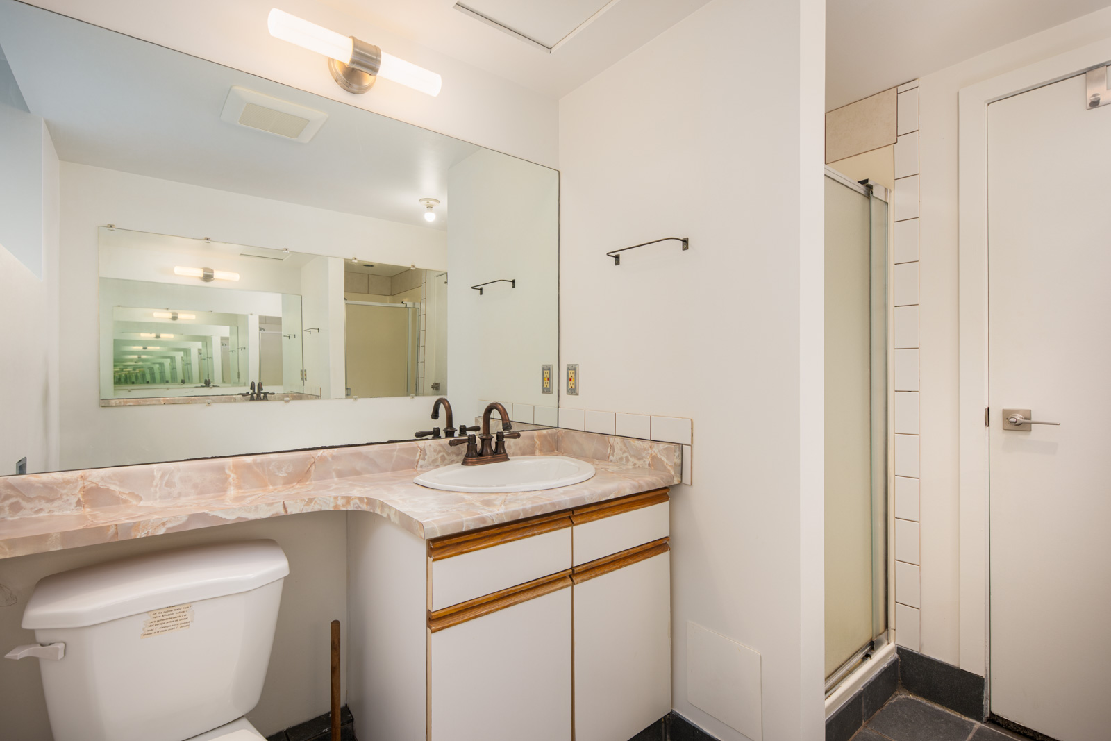 Bathroom with a marble countertop, sink, mirror, toilet, shower with glass door, and white walls. The large mirror reflects the room, creating a repeating effect.