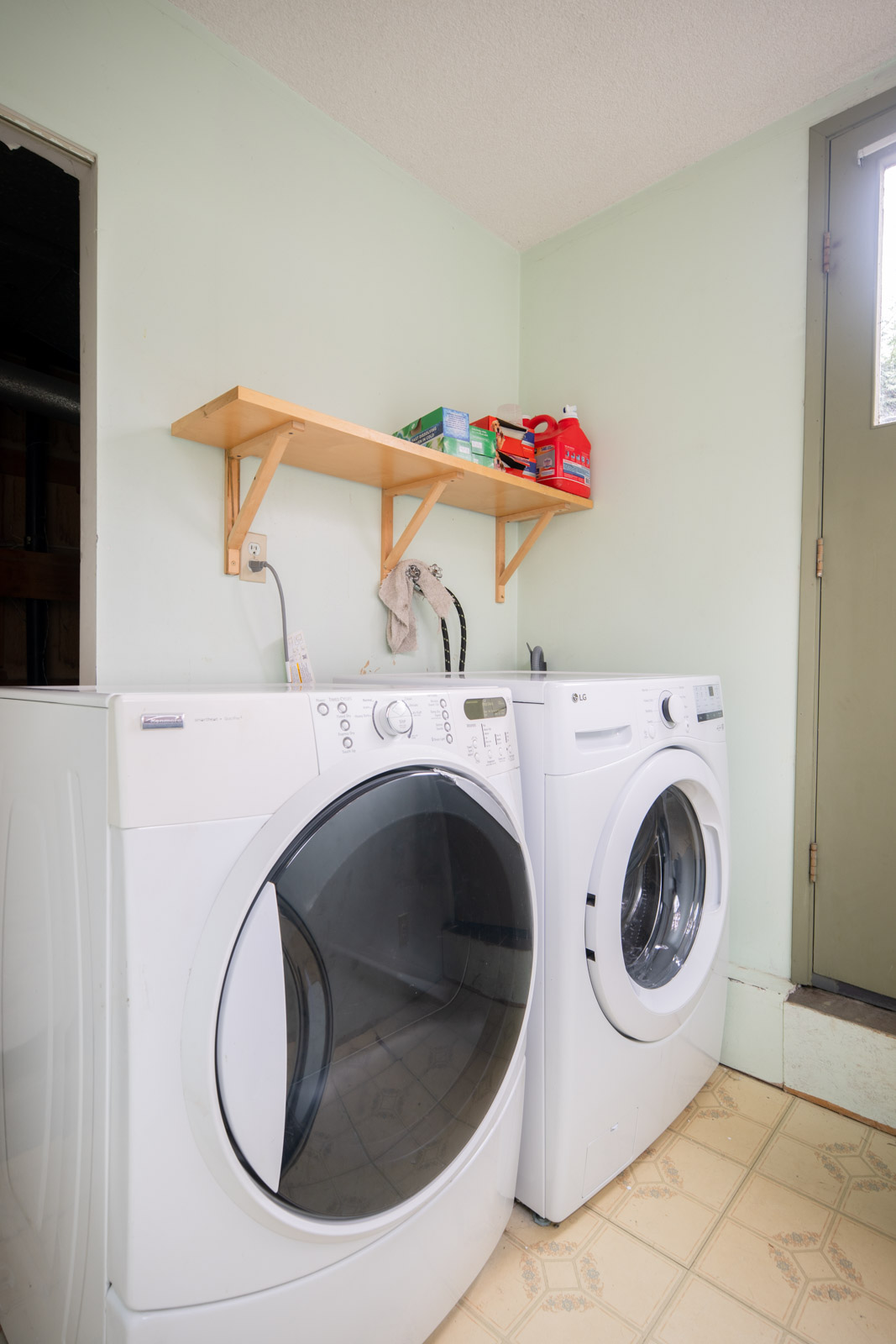 A laundry room with a front-loading washer and dryer, a wooden wall shelf holding detergents above, and a door to the right.