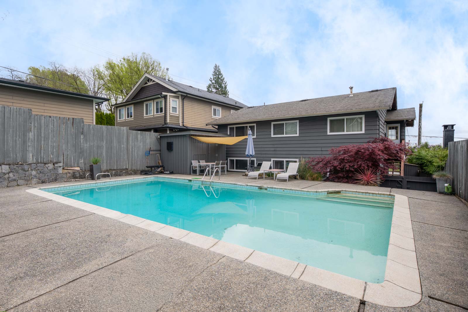 Backyard with an in-ground swimming pool, patio seating, umbrella, and a two-story house with gray siding and a wooden fence.