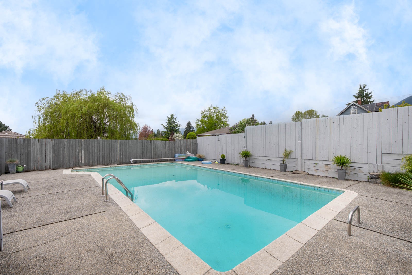 Rectangular outdoor swimming pool with clear water, surrounded by a concrete deck, lounge chairs, potted plants, and enclosed by a tall wooden fence under a blue sky.