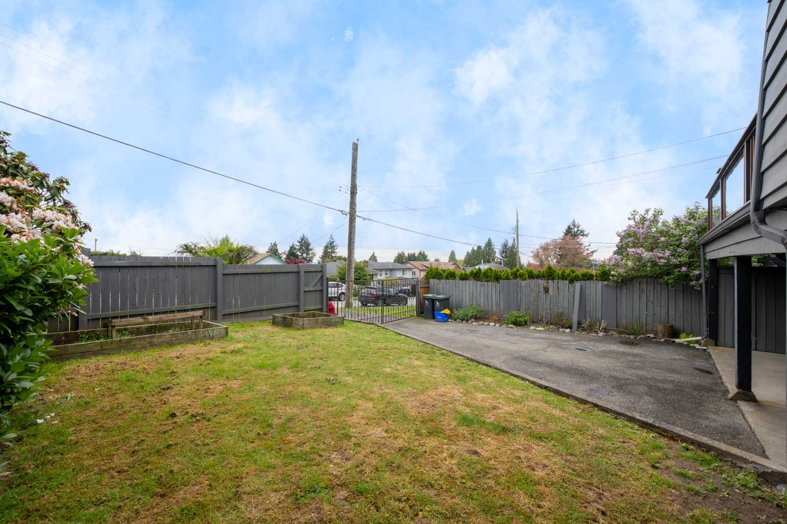 Fenced backyard with a grassy lawn, raised garden beds, concrete driveway, and view of neighboring houses under a partly cloudy sky.