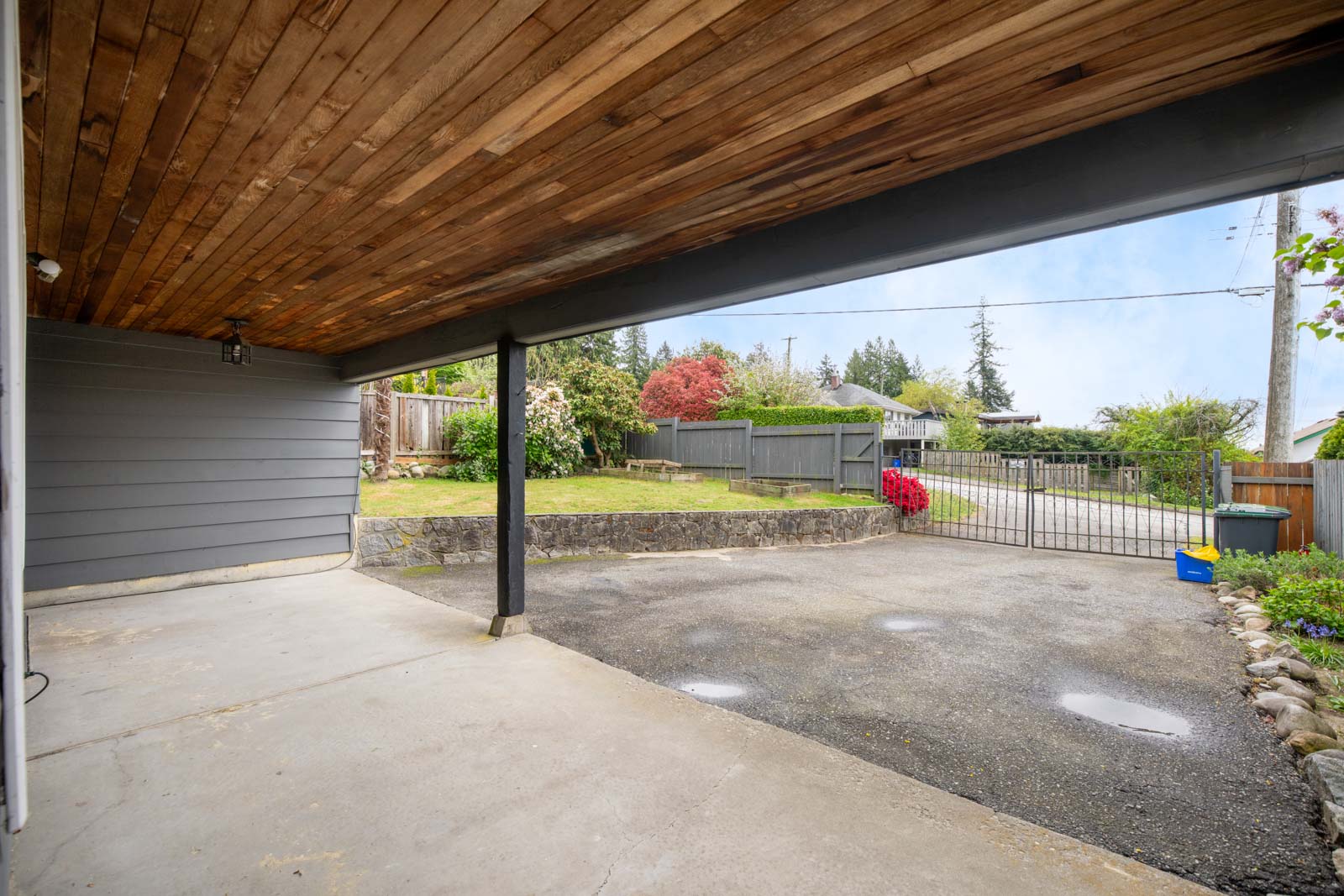 Covered carport with a wooden ceiling, concrete floor, and view of a paved driveway, gated entrance, and a yard with blooming shrubs and a stone retaining wall.