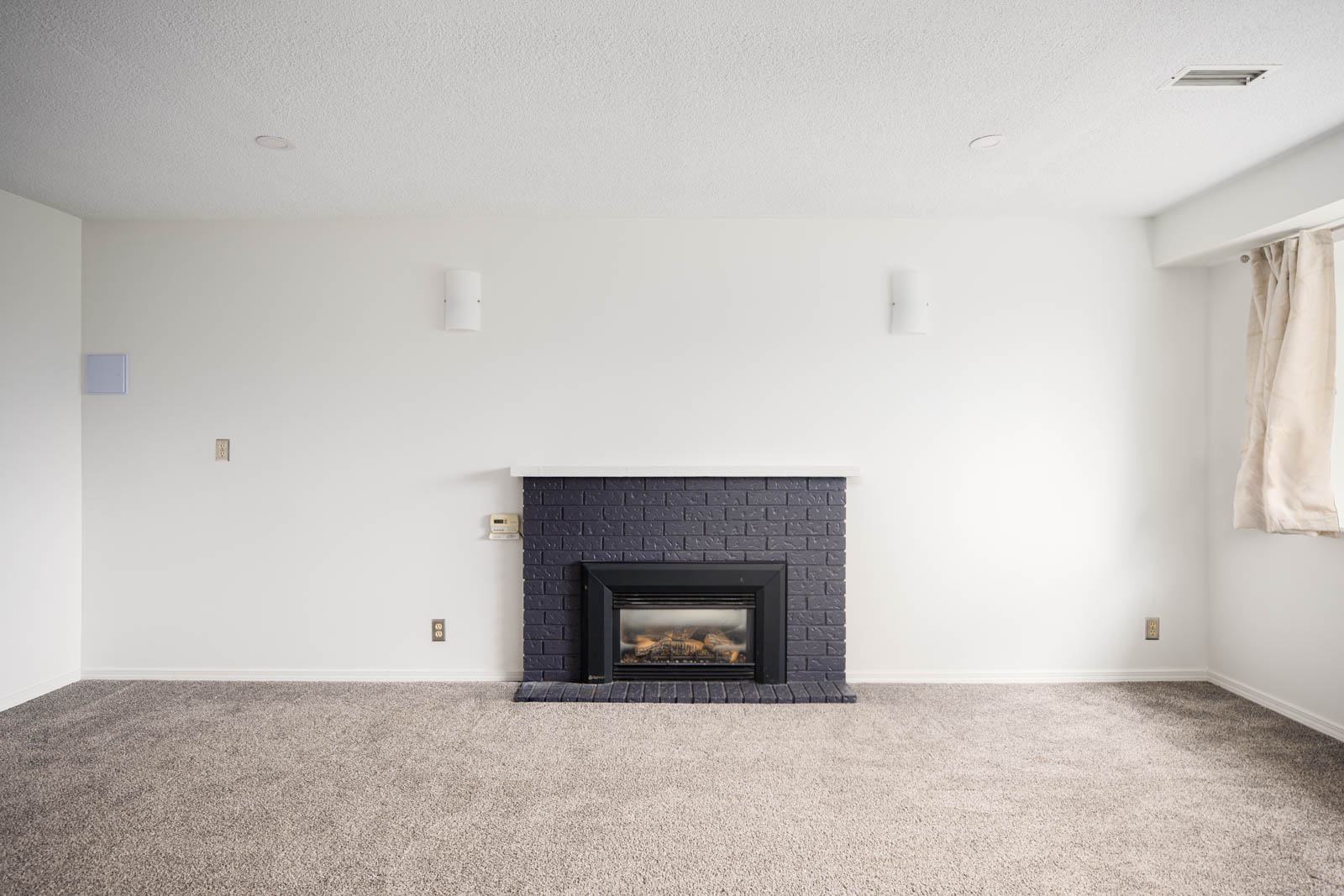 A minimalist living room with white walls, beige carpet, a black brick fireplace, and a window with beige curtains on the right.