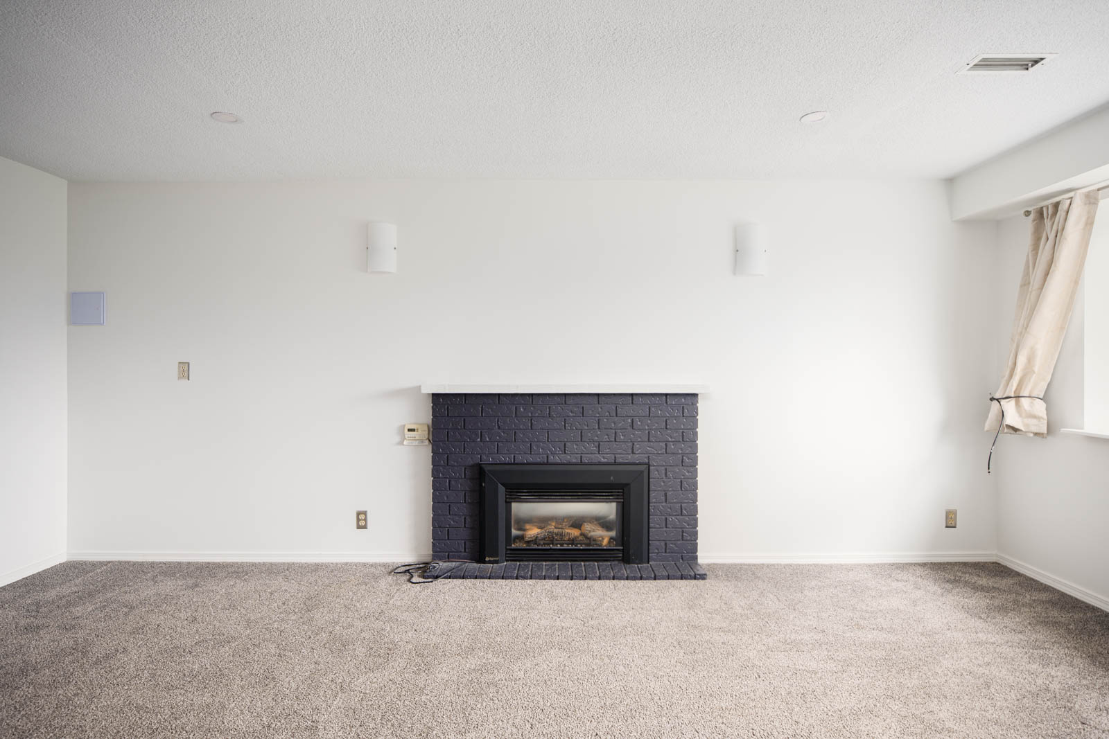 A minimalist living room with white walls, gray carpet, a black brick fireplace, and a window with beige curtains on the right side.