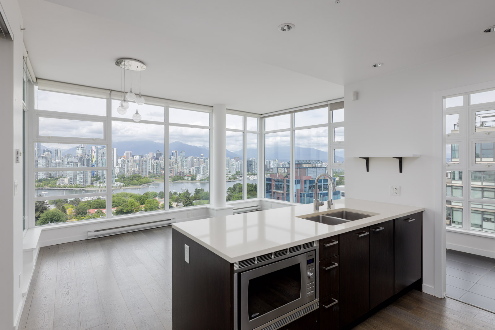 Modern apartment interior with dark wood kitchen cabinets, white countertops, and large windows offering a cityscape and mountain view.