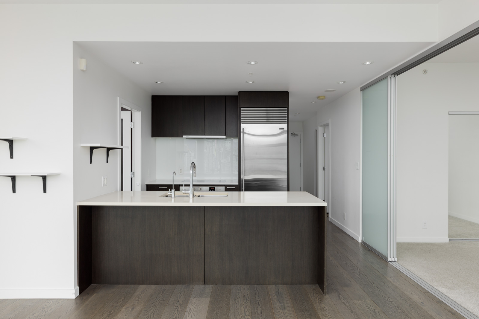 Modern kitchen with dark wood cabinets, stainless steel appliances, a white countertop island, and open shelves, adjacent to a room with carpet visible through sliding glass doors.