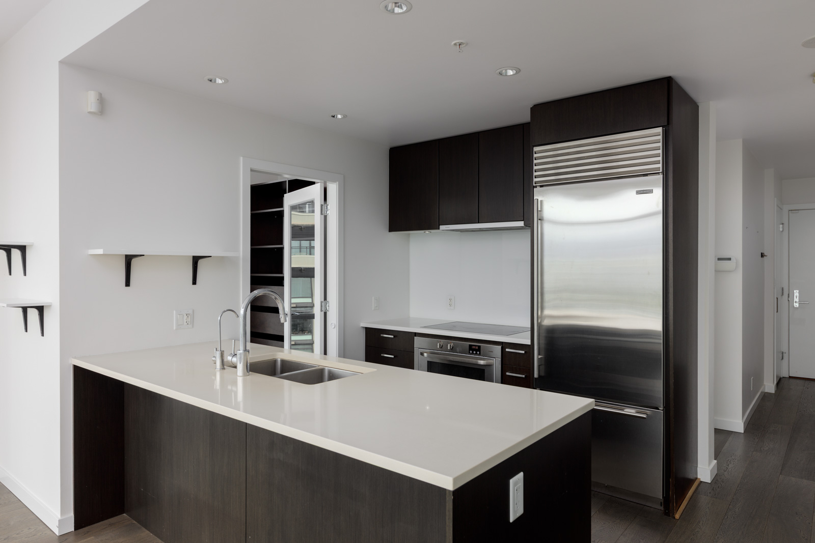 Modern kitchen with dark cabinets, stainless steel appliances, a large refrigerator, white countertops, and an island sink, adjacent to a glass door and minimalist shelving.