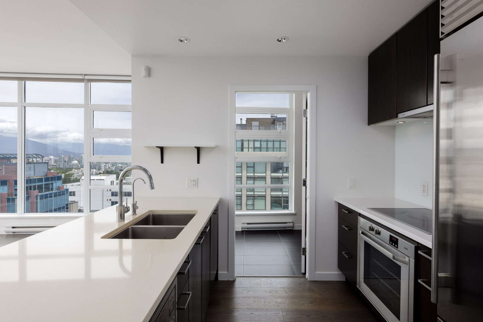 Modern kitchen with white countertops, stainless steel appliances, and large windows overlooking city buildings and mountains in the background.