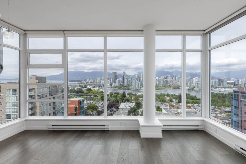 Corner room with large floor-to-ceiling windows overlooking a city skyline, mountains, and water, with gray wood floors and a central white column.