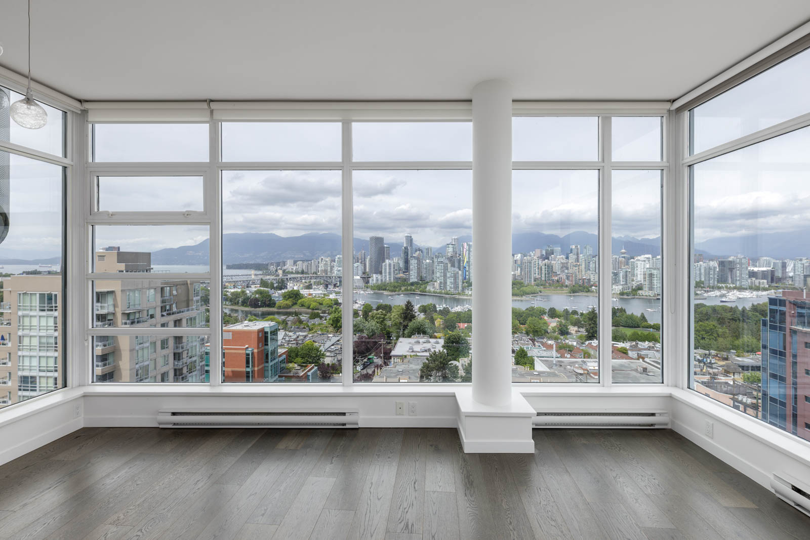 Corner room with large floor-to-ceiling windows overlooking a city skyline, mountains, and water, with gray wood floors and a central white column.