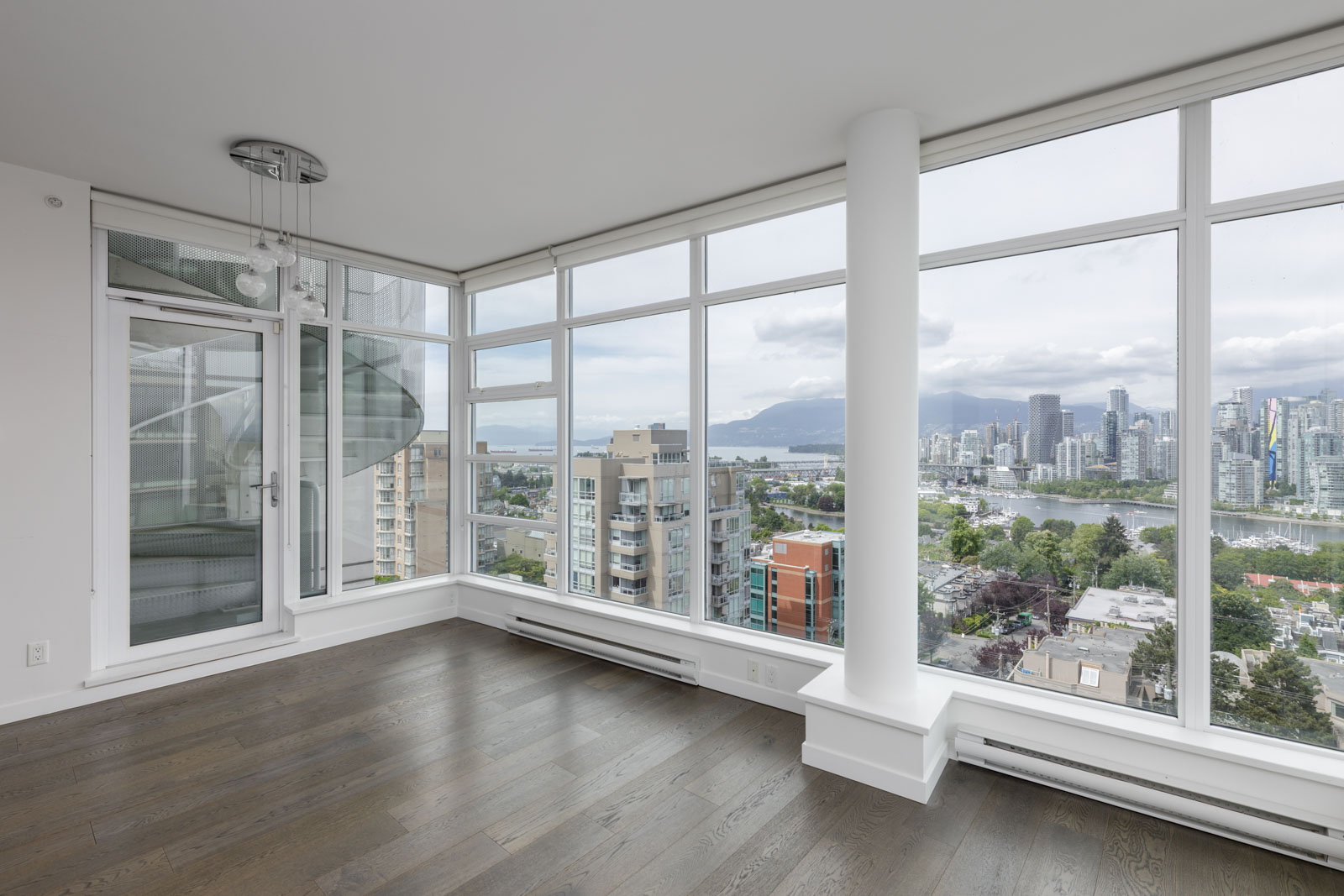 Corner room with large floor-to-ceiling windows, wood flooring, and a view of city buildings, water, and mountains under a cloudy sky.