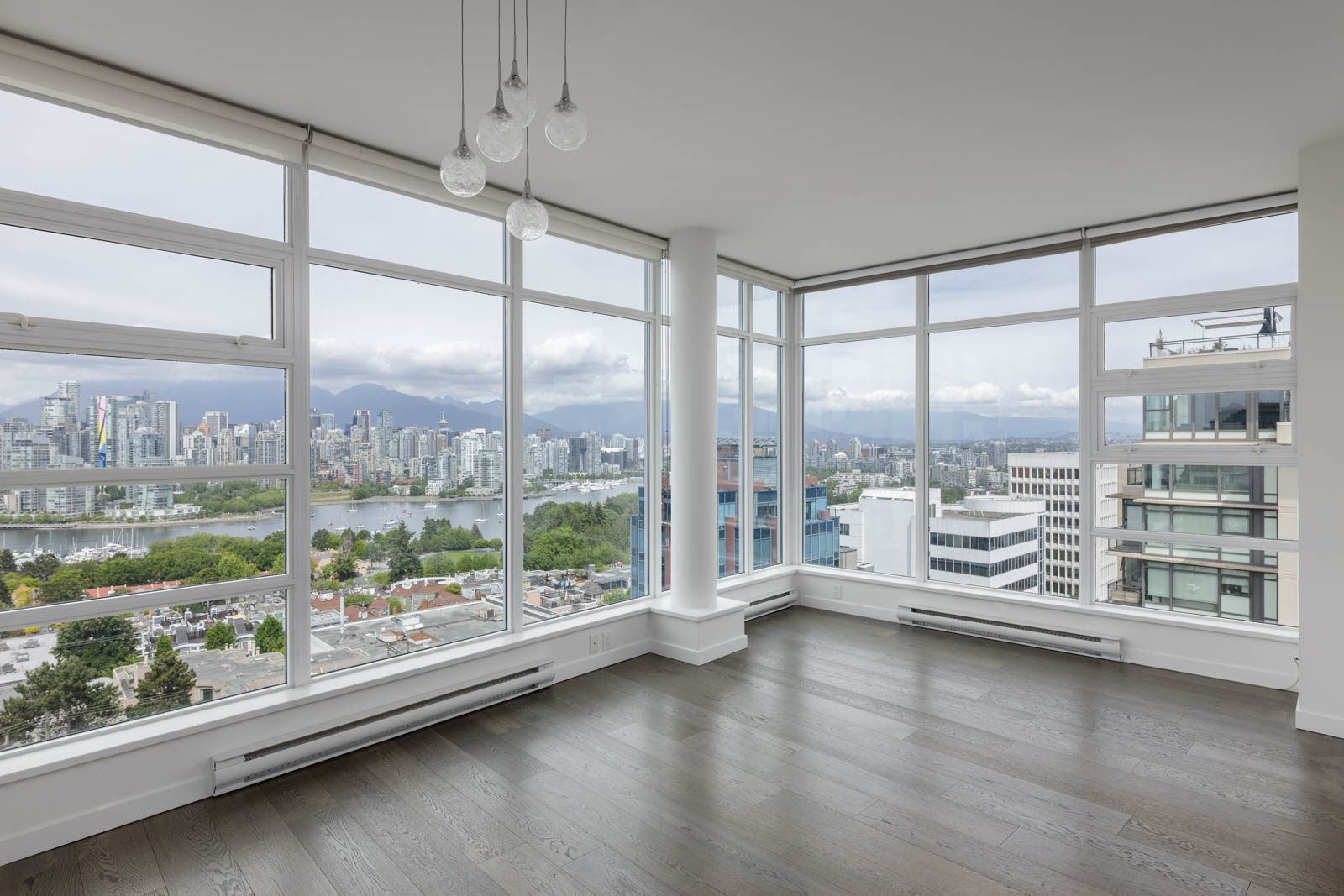 Empty modern apartment with large floor-to-ceiling windows, dark wood floors, and a cityscape view with mountains in the background.
