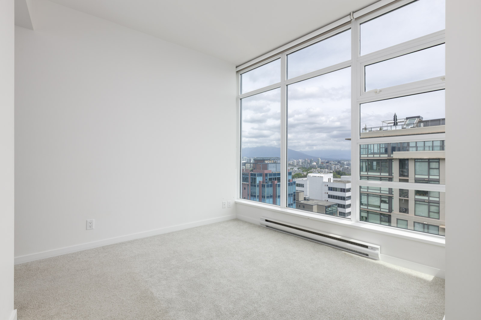 Empty room with white walls, carpeted floor, and large floor-to-ceiling windows offering a city view with neighboring buildings and cloudy sky.