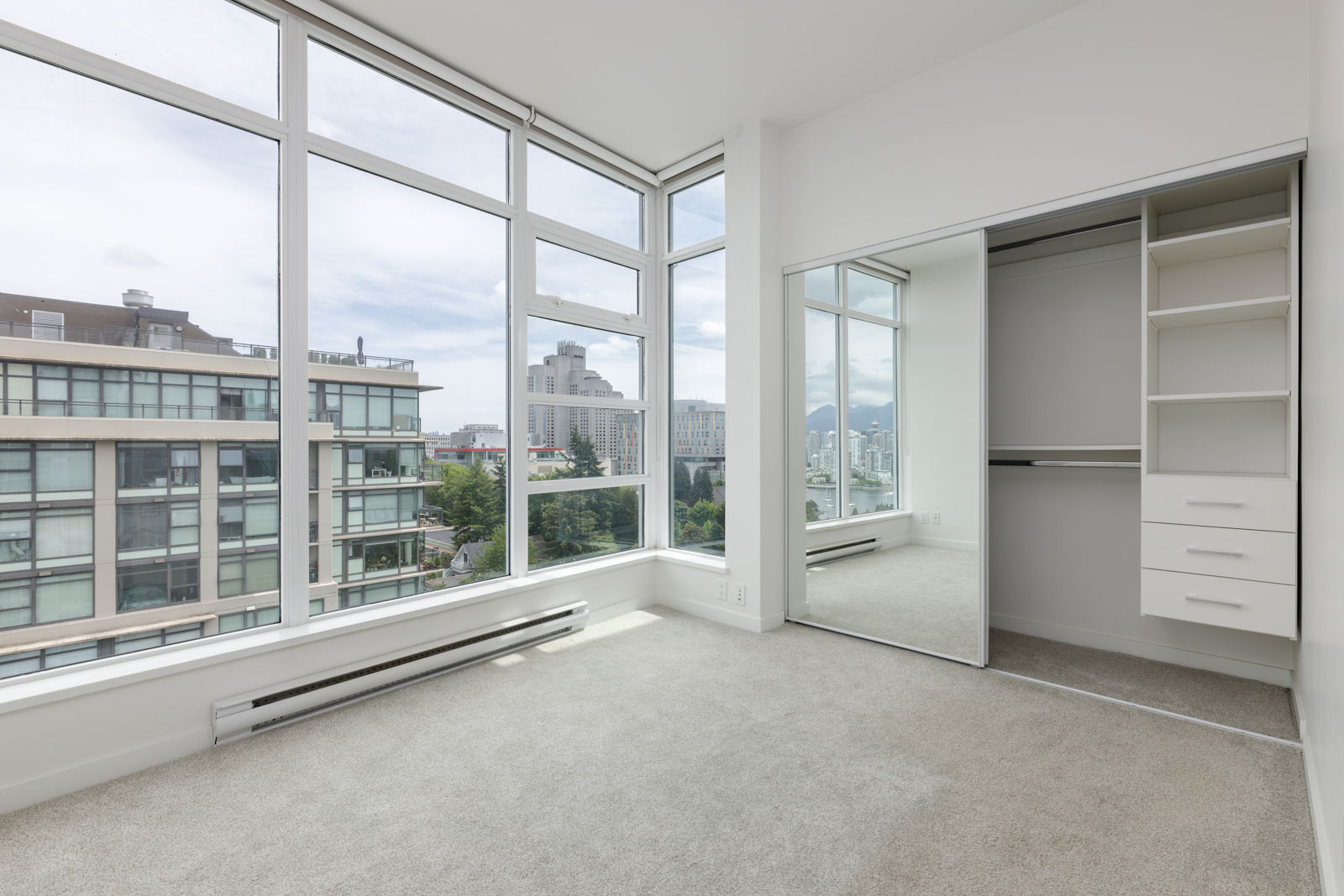 Empty bedroom with large floor-to-ceiling windows, a mirrored sliding closet door, built-in shelves, and a view of city buildings outside.