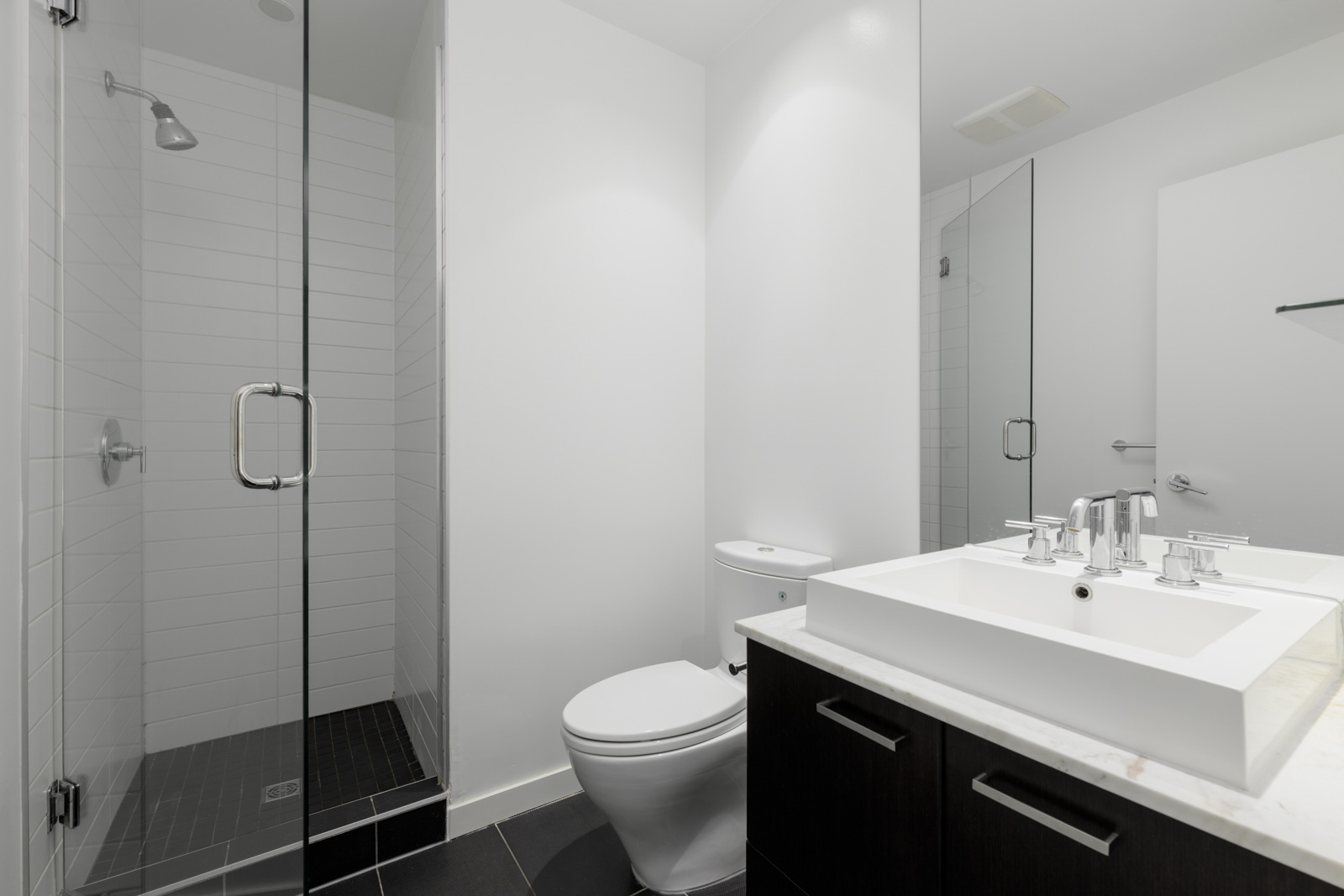 Modern bathroom with a glass-enclosed shower, white toilet, dark vanity with a white countertop, and a large mirror.